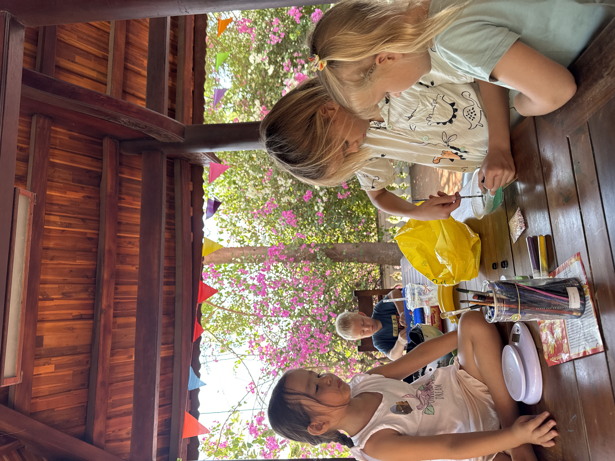 This outdoor scene captures three children engaged in a creative activity at a wooden table, likely in a covered patio or open-air area in Mũi Né, Vietnam. The wooden structure features dark beams and a lighter plank ceiling/wall, providing shade. Vibrant green foliage and clusters of bright pink flowers (possibly bougainvillea) are visible in the background, along with colorful triangular bunting flags, suggesting a cheerful atmosphere.
In the foreground, a young girl with dark hair, tied in small braids, is seated, looking down at the table. She wears a light-colored t-shirt with "DREAM" printed on it. At the center of the image, two blonde-haired children are focused on their crafts. One girl, with her hair partially tied back with a colorful scrunchie, wears a light shirt adorned with dinosaur patterns and is actively painting or crafting with a small brush and a container of green substance. Another blonde child is partially visible next to her. Further in the background, a boy with light hair is also seated at a table, wearing a dark t-shirt with the partial text "LIFE'S A...".
The table is laden with art supplies, including a clear container holding numerous colored pencils and brushes (with "VOLUME 7.1L" visible on its side), small packets of beads or glitter, and a plastic cup with a straw. A yellow plastic bag and a small purple and white weighing scale are also on the table. The bright lighting suggests it is daytime, likely midday or afternoon, under sunny weather conditions. The children appear focused and engrossed in their activity.