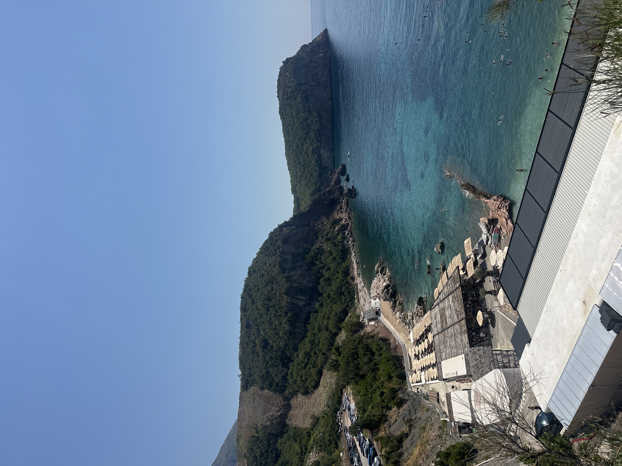 The image captures a panoramic view of a coastal area in Sveti Stefan, Montenegro, on a clear, sunny day. The deep blue sea meets a rocky shoreline, with a lush green mountainous landscape rising in the background.
In the foreground, a modern beach club facility is visible, featuring a wooden-roofed structure with multiple seating areas and umbrellas. Several beach chairs are arranged on the sand and around the club. To the left of the beach club, a road winds up the side of the mountain, with a parking lot filled with numerous cars.
The sea itself is a vibrant turquoise color, becoming deeper blue further out. The water is populated by several individuals swimming and enjoying the ocean. A floating rope barrier marks a designated swimming area in the water.
The architecture of the beach club is contemporary, with clean lines and natural materials like wood and stone. The surrounding environment is characterized by steep, verdant hillsides and a rugged coastline. The overall impression is one of a popular tourist destination offering relaxation and recreation by the sea.