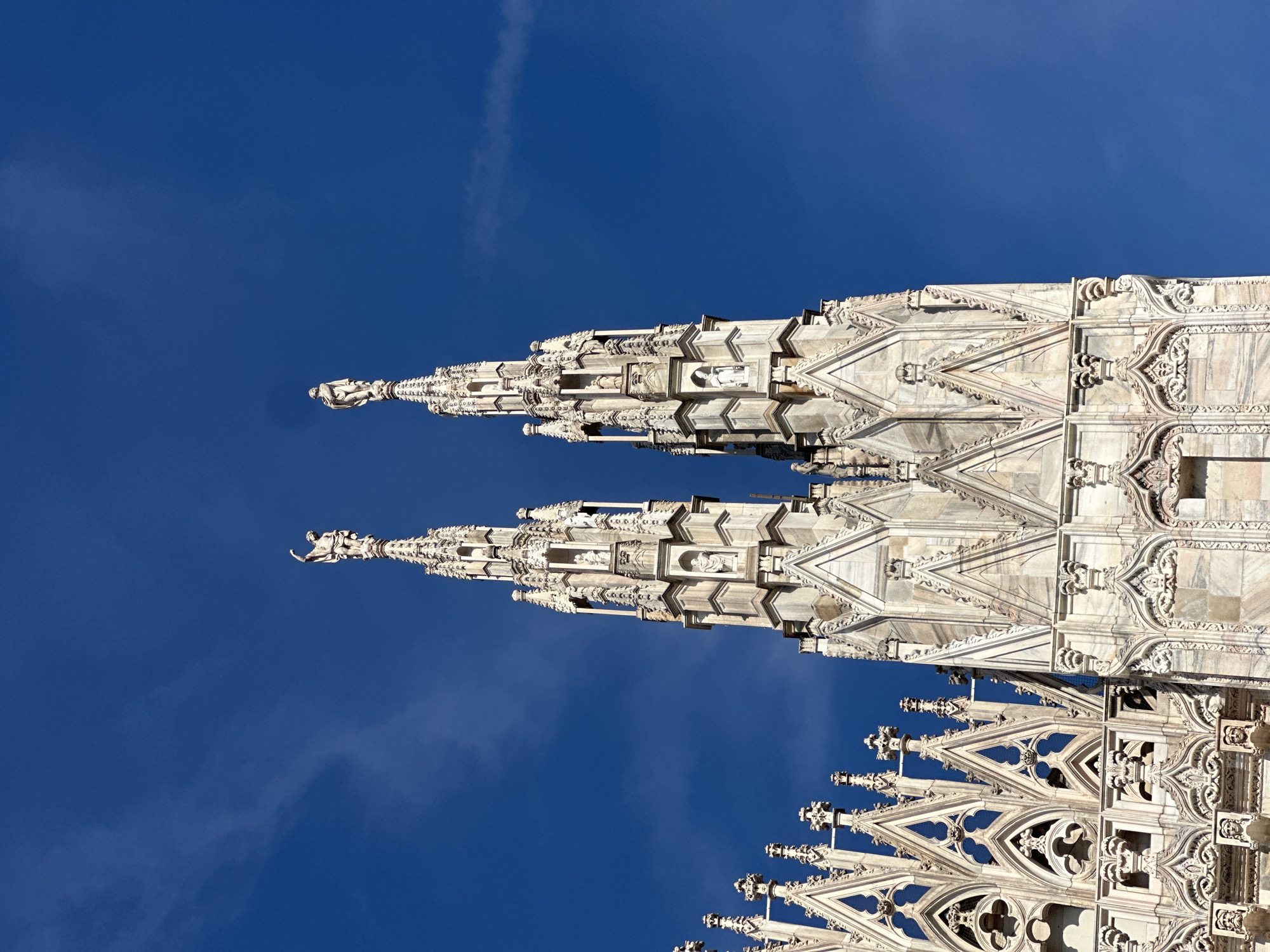 The image shows a low-angle, full-shot of two spires of the Duomo di Milano (Milan Cathedral) against a clear, vibrant blue sky. The spires are ornately decorated with Gothic architecture, featuring intricate stone carvings, small statues within niches, and pointed pinnacles. Atop each of the two prominent spires, there is a statue of a figure, one with its arm raised. The stone of the cathedral appears to be a light-colored marble. The detailed stonework suggests a significant architectural landmark. The sky is largely cloudless, with a hint of wispy clouds. The composition focuses on the height and grandeur of the spires.