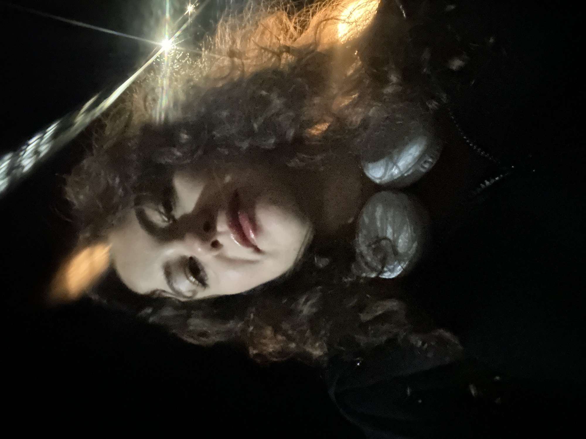 The image is a low-angle, dark photograph of a person with curly hair, appearing to be a selfie. The individual has fair skin and is wearing what appears to be two circular metallic earrings or other items near their ears. The background is mostly black, but there is some light reflecting off objects in the upper left, creating a bright flare. There is no clear indication of the location being Budva, Montenegro, or any other specific location.