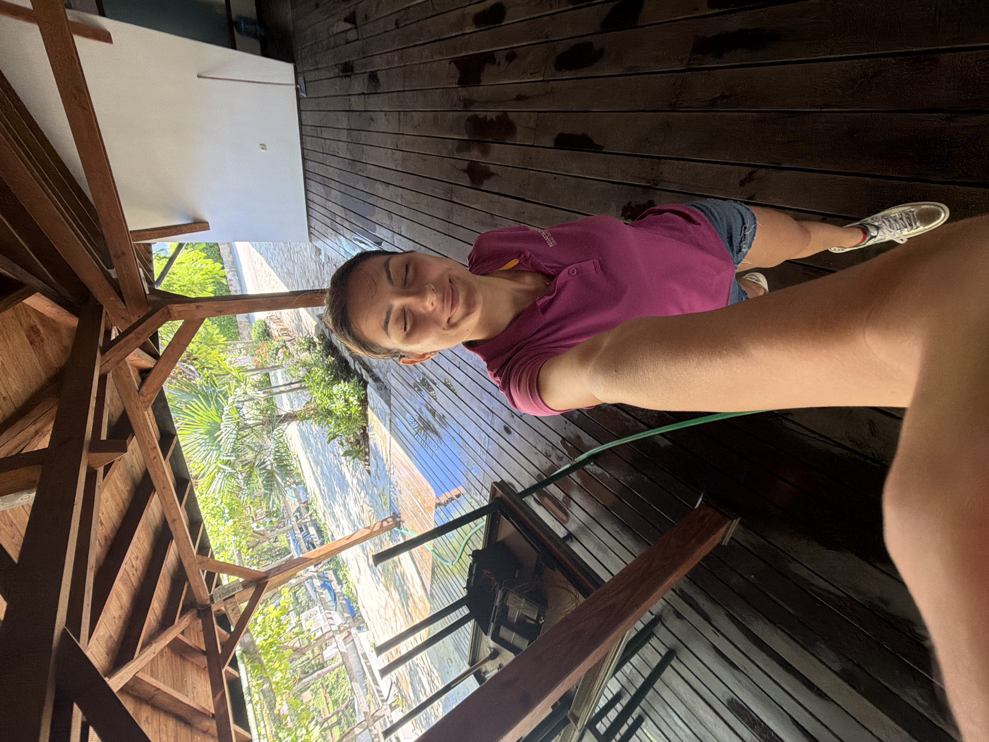 A woman is taking a selfie under a wooden gazebo. She is wearing a purple polo shirt and denim shorts, and is smiling with her eyes closed. The wooden deck of the gazebo is wet, suggesting it has recently rained or been cleaned. Outdoor foliage and trees are visible beyond the gazebo. To the left, a table and chairs are overturned. The overall scene appears to be a residential or commercial outdoor space, possibly near a garden or park.