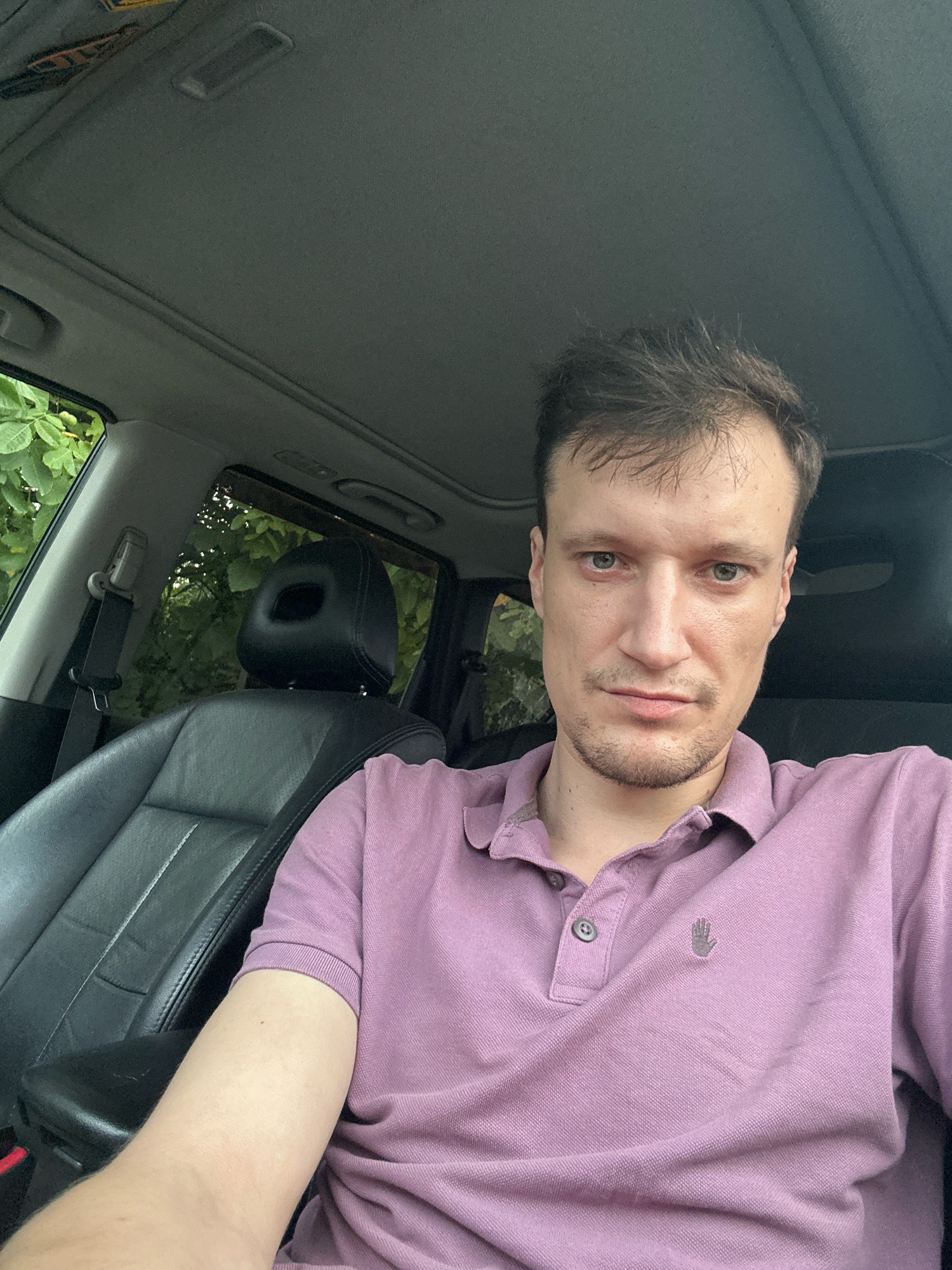 A man with light skin, brown hair, and blue eyes is sitting in the driver's seat of a car. He is wearing a pink polo shirt with a small grey hand logo on the left breast. The car interior is visible, including black leather seats, a sunroof, and a seatbelt. Outside the car, green foliage is visible through the windows. The image appears to be a selfie taken in daylight. The location is specified as Shatava, Ukraine.