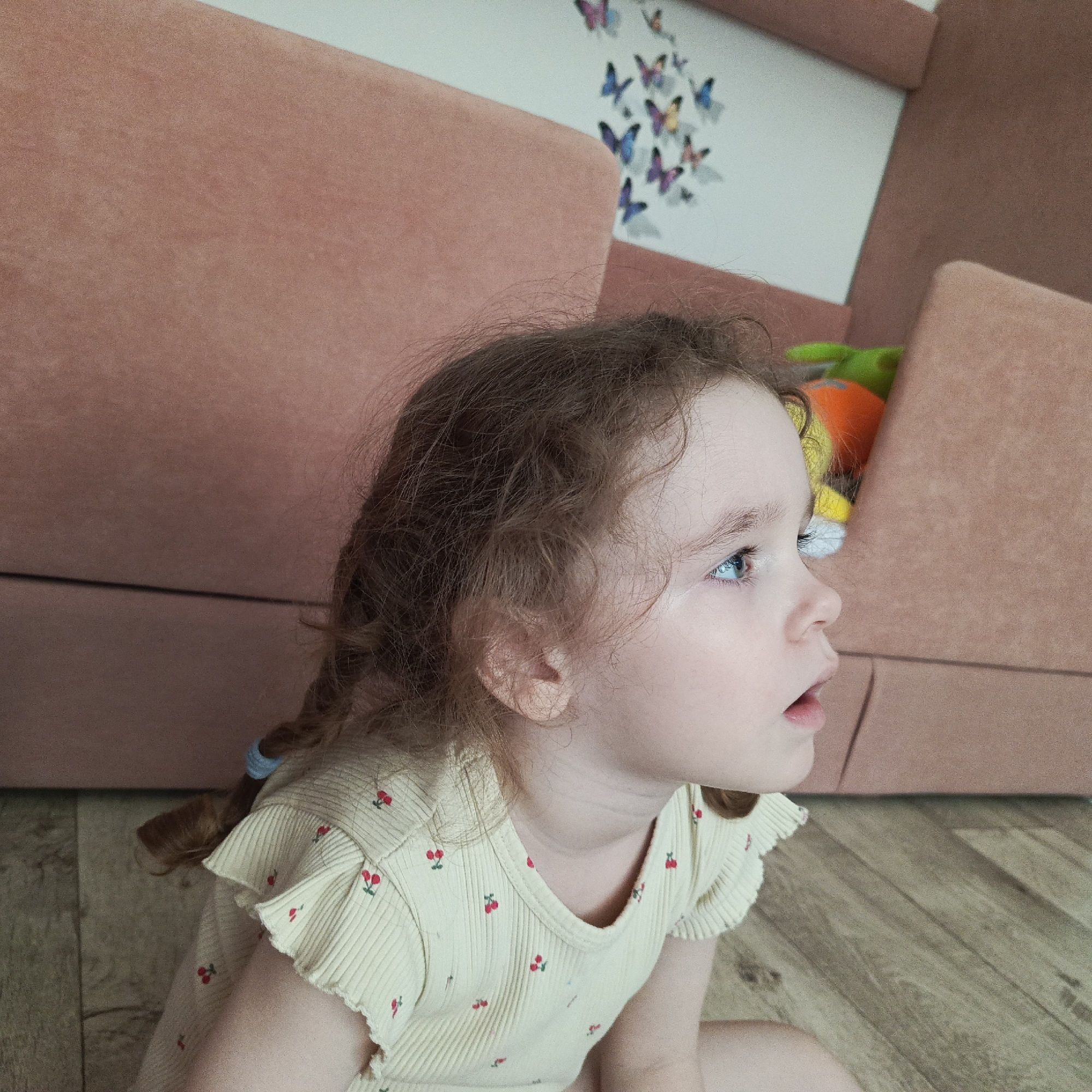 A young child with curly, reddish-brown hair sits on a light wood-grain floor in Марковский, Russia. The child is dressed in a light yellow shirt adorned with small red cherry patterns and small ruffles on the sleeves. Her hair is partially braided and secured with a light blue hair tie. She is looking to the right, with an expression of surprise or interest, her mouth slightly open. Behind her, part of a dusty rose-colored couch is visible. The wall behind the couch is white with several butterfly decorations. A toy in shades of green, orange, and yellow is peeking over the top of the couch.