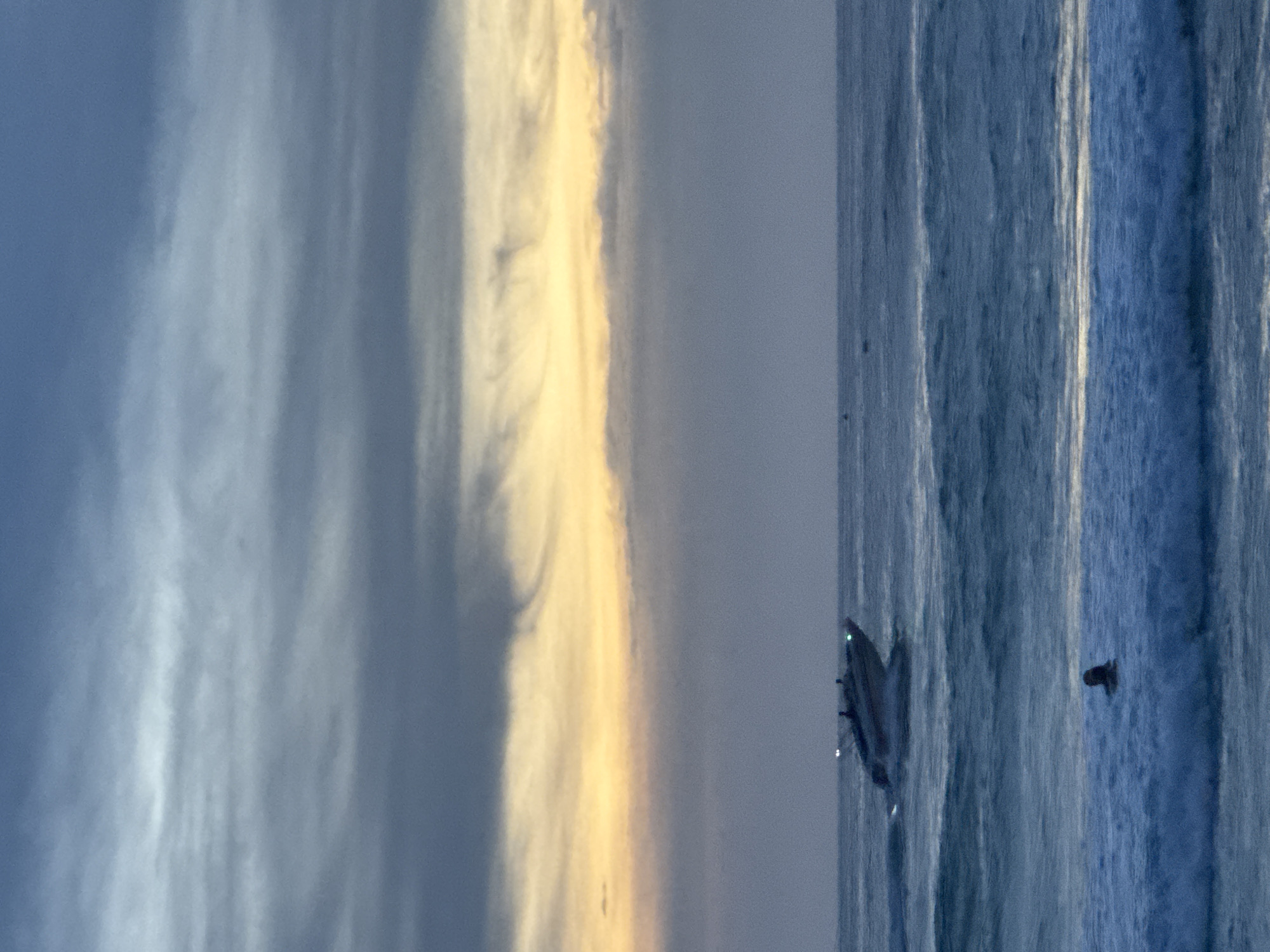 In Karon, Thailand, at dusk, a fast-moving white motorboat with at least two occupants heads toward the right of the frame, leaving a white wake on the choppy blue sea. A single person is visible in the surf in the foreground, appearing to swim or float in the water. The sky is a gradient of muted blues and grays, with a band of soft yellow and orange near the horizon, suggesting a sunset. Wispy clouds stretch horizontally across the sky.
