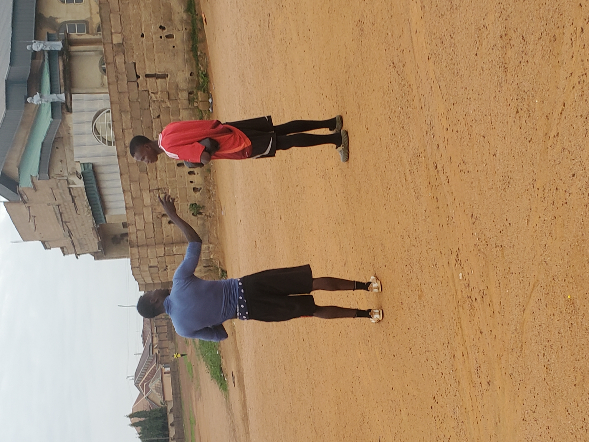 Two young men stand on a dirt road in Jos, Nigeria. The man closer to the camera wears a blue long-sleeved shirt and black shorts with a polka dot trim. His left arm is raised. The other man wears a red shirt and black shorts. Buildings are visible in the background. The sky is overcast.