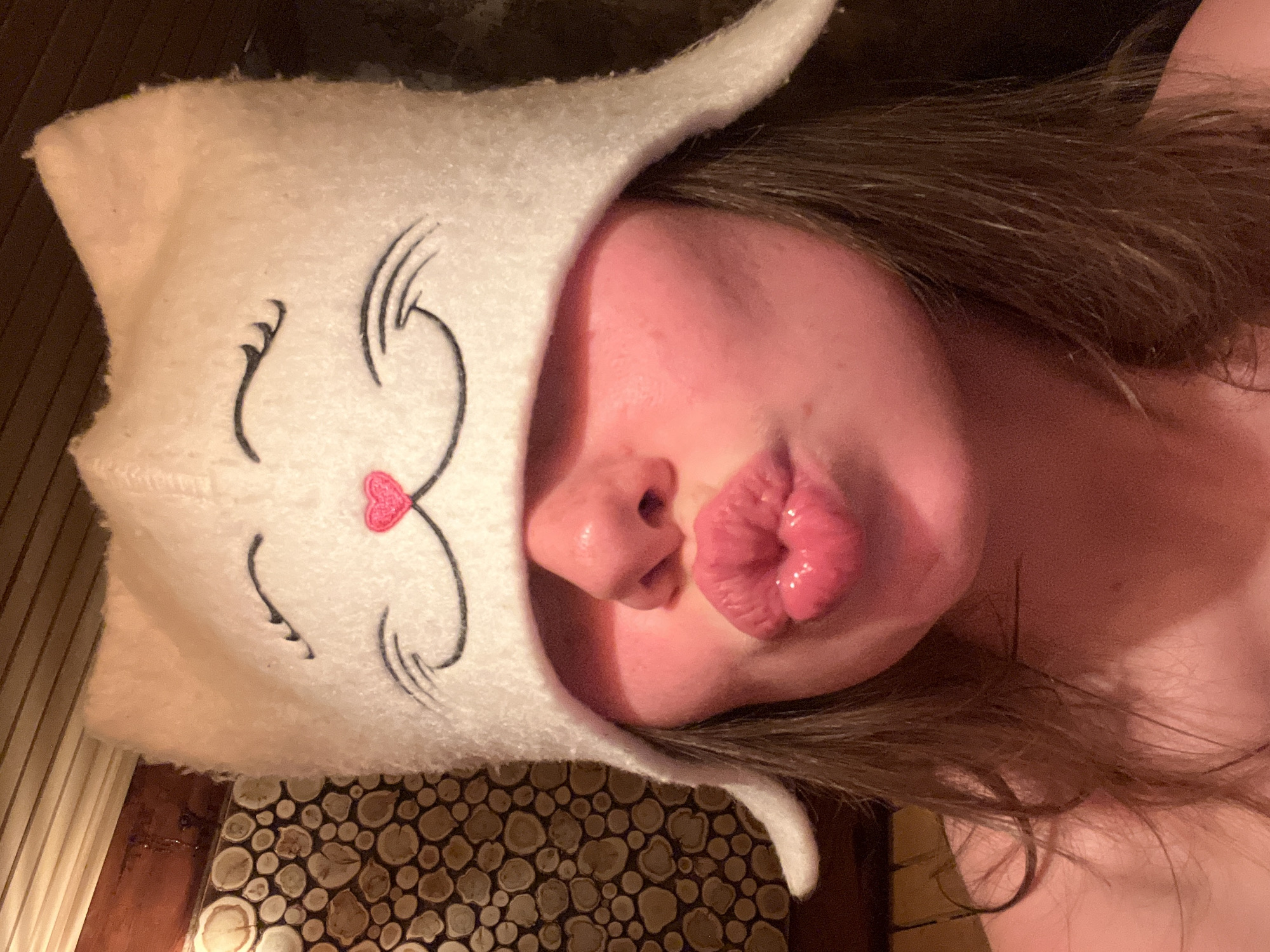 A selfie depicts a person wearing a white cat-eared hat with a stitched-on cartoon cat face, puckering their lips toward the camera. The person has shoulder-length brown hair. The background shows a wooden surface with a circular pattern. The location is stated as Chaikovsky, Russia.
