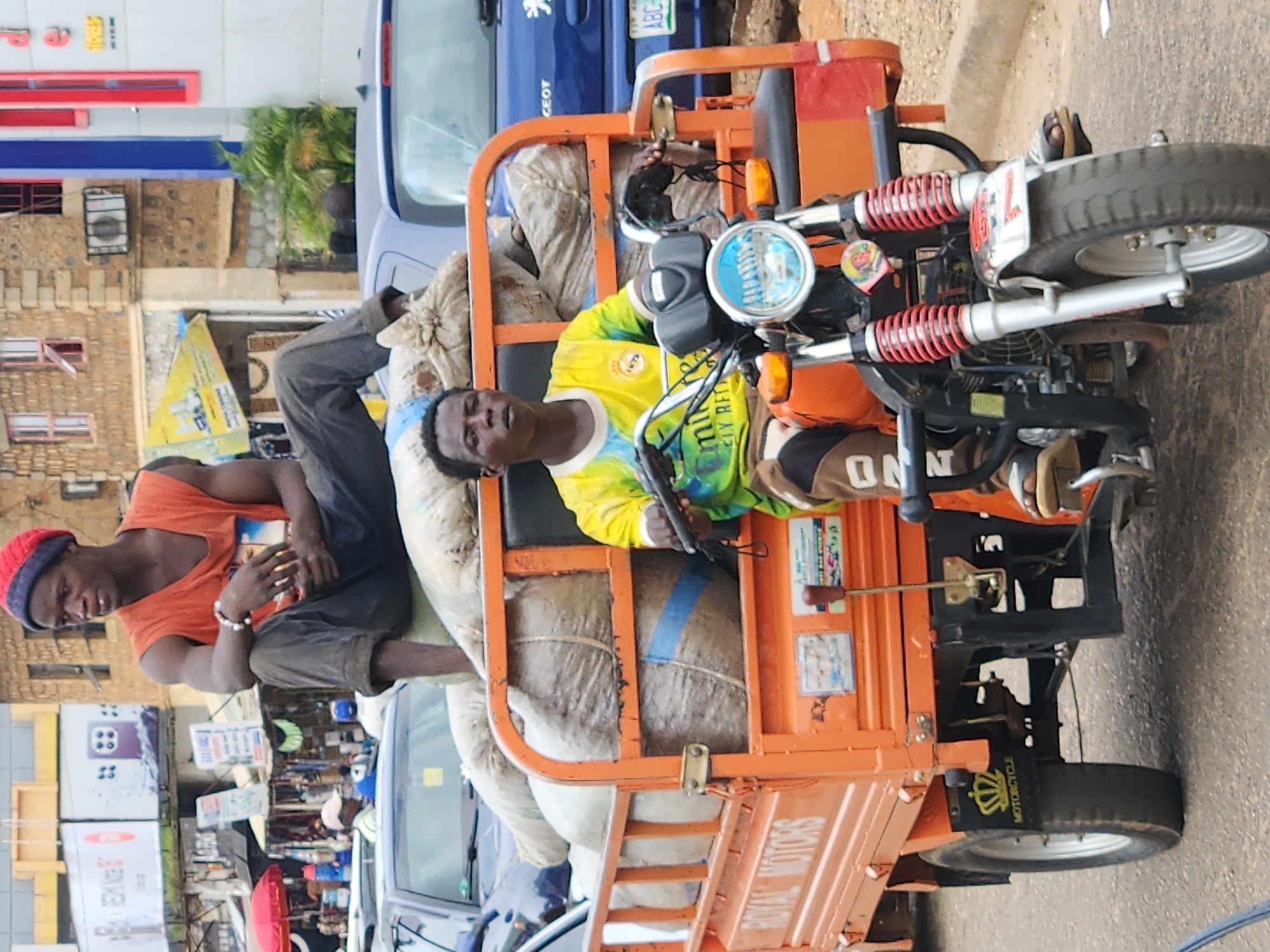 This is a medium shot, eye-level, outdoor photograph taken in Jos, Nigeria. The scene depicts two young men and a three-wheeled orange transport vehicle filled with bagged goods. The vehicle is parked on an asphalt road with a curb and sidewalk visible. In the background, there are buildings, signs, and other vehicles, suggesting an urban environment.
The man in the foreground is seated on the driver's seat of the vehicle, wearing a yellow and green t-shirt and brown pants. He is looking directly at the camera. The man in the background is seated on top of the bagged goods, wearing an orange tank top and dark pants, with a red and blue beanie. He is looking to the side.
The three-wheeled vehicle has "ROYAL MOTORS" written in white on its side. The cargo area is filled with numerous large, light-colored bags, some secured with blue straps. The vehicle's front features a single wheel and a headlight.
The background includes a multi-story building with a stone facade, red window frames, and blue architectural elements. A large billboard with text and graphics is also visible, along with other shops and market stalls. Several cars are present, including a blue car behind the transport vehicle. The lighting suggests it is daytime.