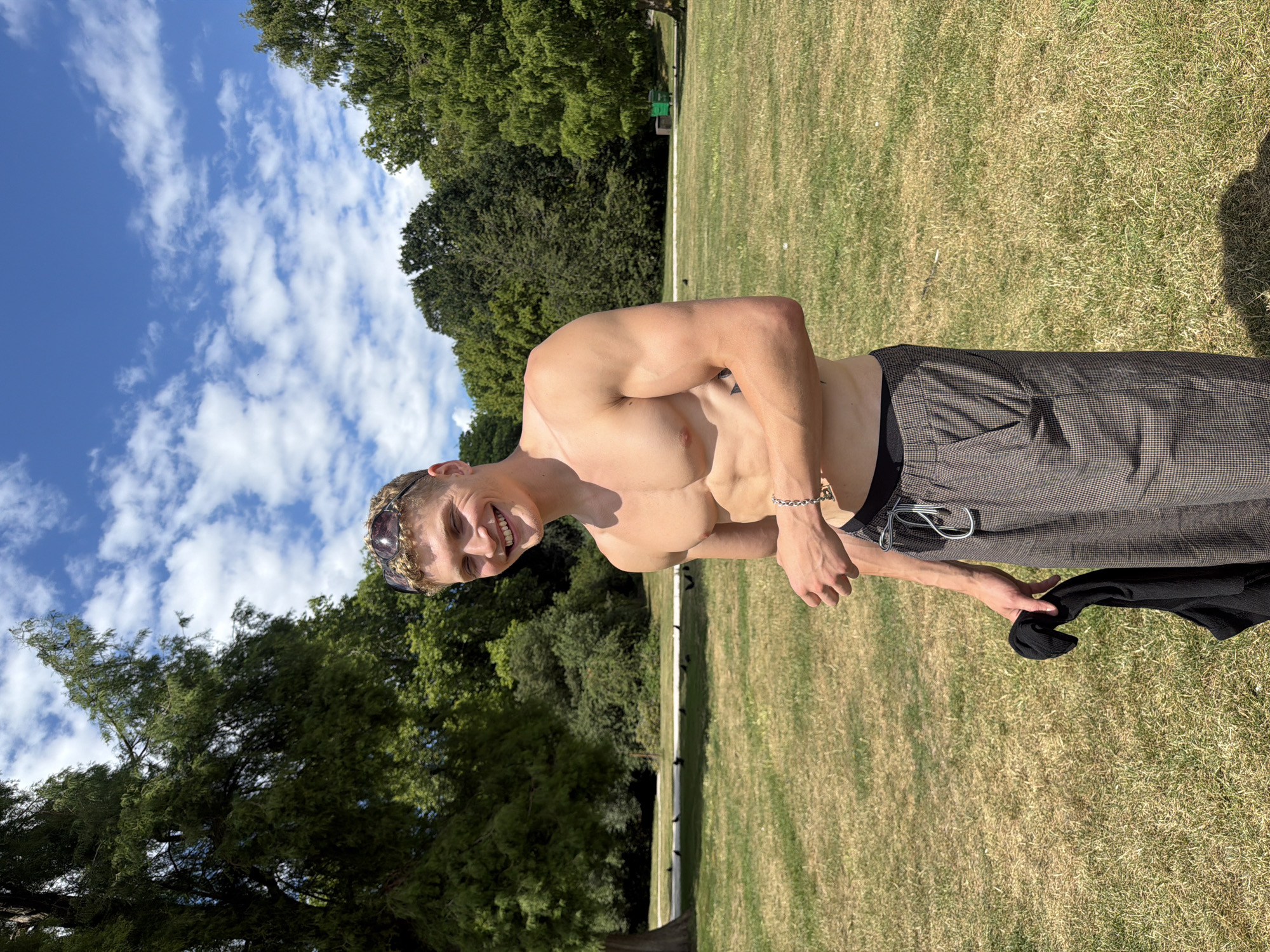 A shirtless man with blonde curly hair and sunglasses perched on his head stands in a park on a sunny day. He is wearing checkered trousers and holding a black garment in his left hand. He has a silver bracelet on his left wrist and a tattoo on his left torso. The park has green grass and trees in the background, with a blue sky and white clouds above. The man is smiling at the camera. The scene appears to be Hyde Park in London, United Kingdom.