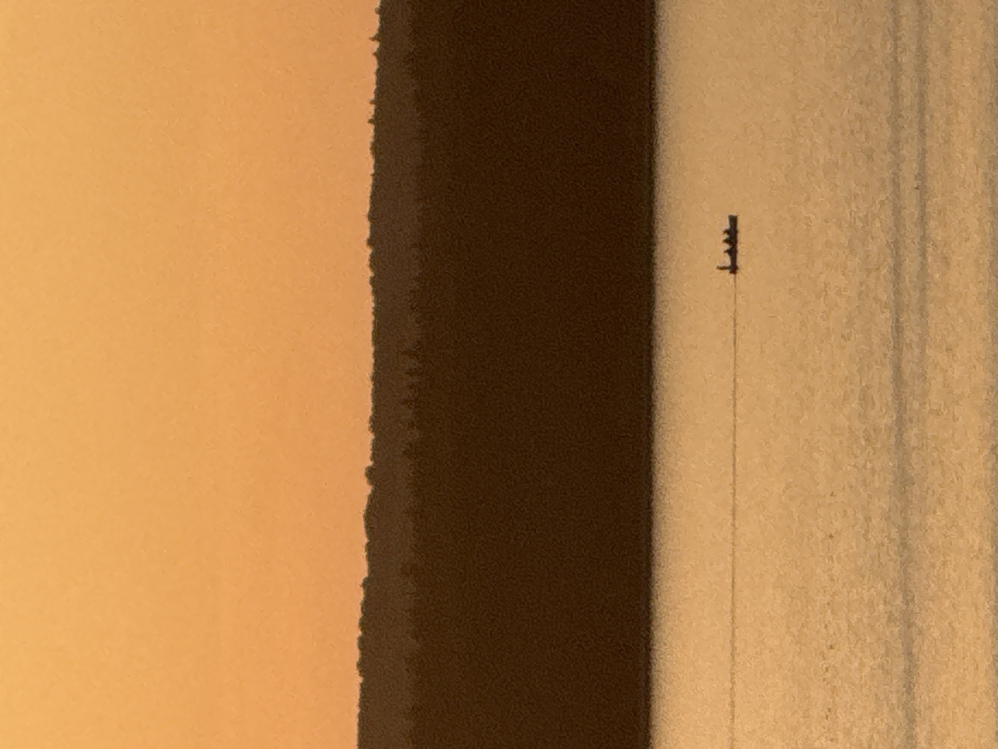 A vertical close-up shot shows a three-signal traffic light mounted on a pole, likely in Votkinsk, Russia. The light is silhouetted against a light tan or beige backdrop. To the left of the traffic light and pole is a dark brown vertical area, potentially representing a building or a shadow. Farther to the left is a light orange area, perhaps a wall or the sky. The overall lighting suggests a sunset or sunrise, with warm tones dominating the scene. The image is slightly blurry or grainy.