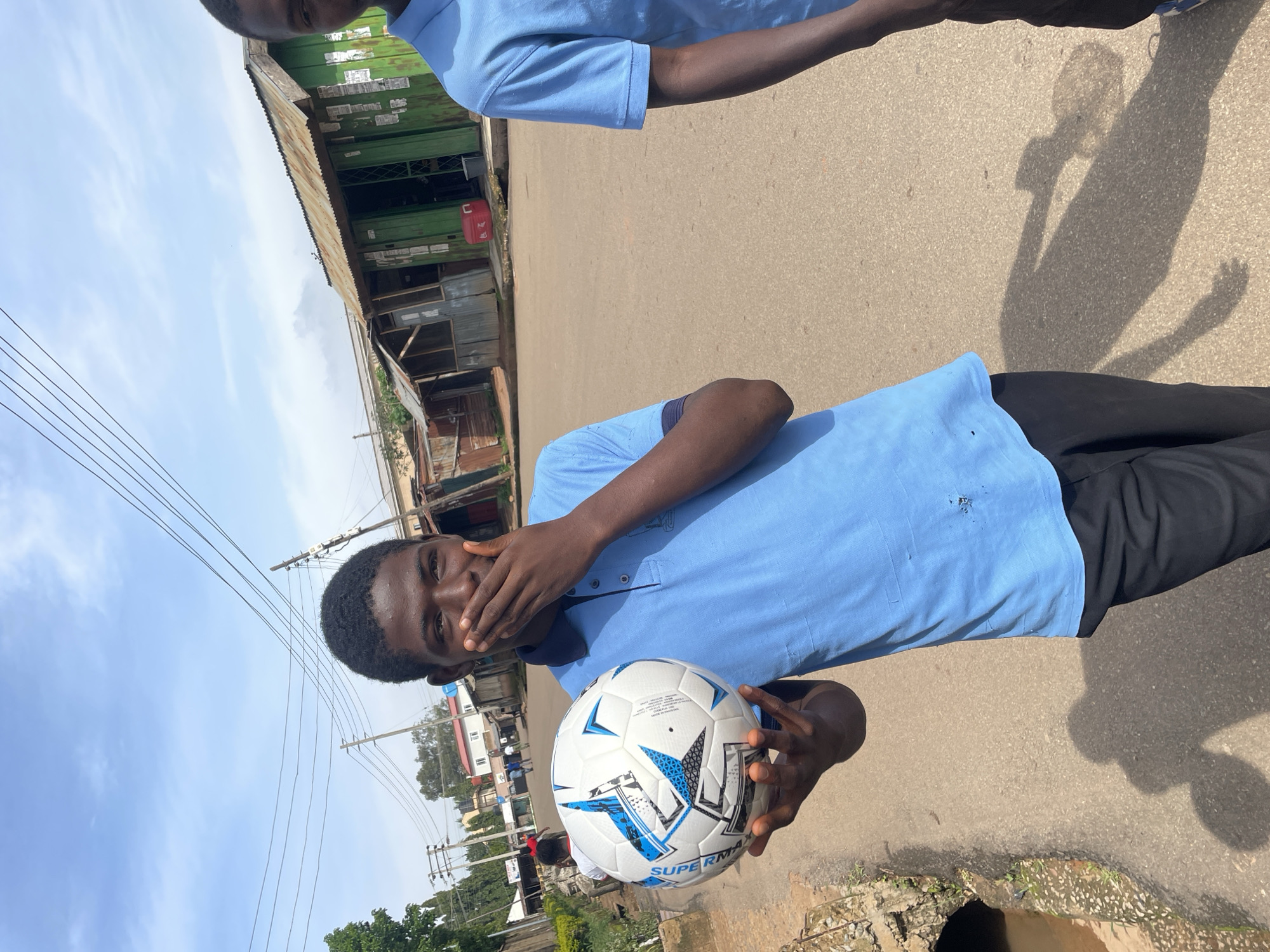 A young Black male, wearing a light blue polo shirt and black pants, stands on a paved road in Gura Top, Nigeria, holding a white and blue soccer ball in his left hand. He is covering his mouth with his right hand, and his head is tilted slightly to his right. The background shows a dusty street with buildings and power lines. On the left side of the image, a drainage ditch runs alongside the road, with some vegetation growing around it. Another Black male, also wearing a light blue polo shirt, is partially visible on the right side of the frame. The sky is clear with a few clouds.
