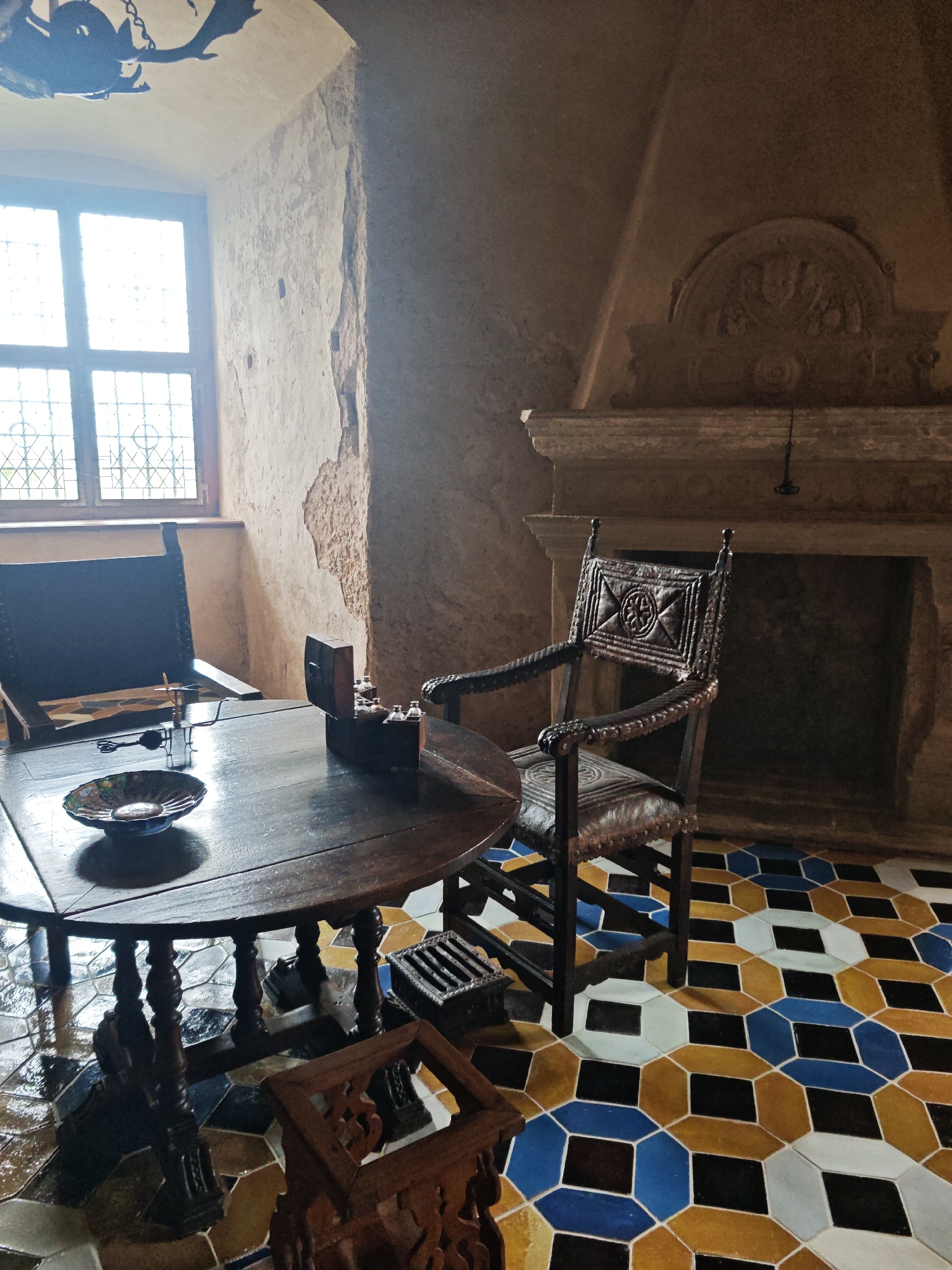 The image depicts an interior scene, likely within the Bauska Castle complex in Bauska, Latvia. The room features a large, rustic wooden table with turned legs, set on a colorful geometric tiled floor in shades of yellow, blue, black, and white. Two chairs are present: one dark leather-upholstered chair with a high back to the left, and an ornate, carved wooden chair with a padded seat and back to the right. On the table, a decorative bowl and a wooden box containing small bottles are visible. A large stone fireplace with a decorative archway is prominent on the right wall, with a faint beam of light illuminating the area above it. A window with leaded glass panes is situated on the left wall, with a dark, decorative hanging light fixture visible in the upper left corner. The walls appear to be rough-plastered, suggesting an aged or historical setting.