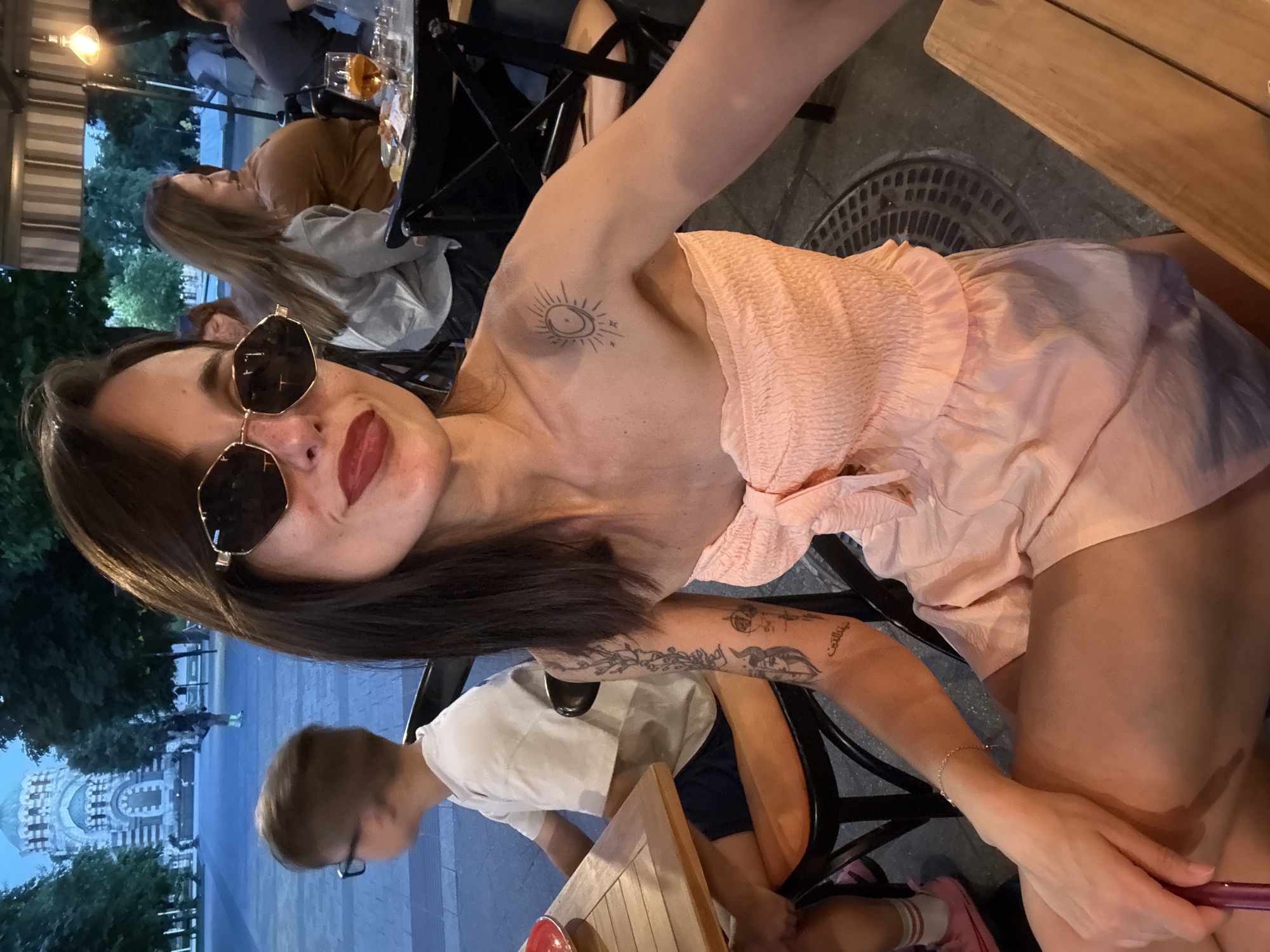 In Pleven, Bulgaria, at an outdoor dining establishment, a woman with dark hair and sunglasses poses for a selfie. She is wearing a strapless peach-colored smocked dress. A small tattoo of a sun and moon is visible on her right shoulder. To her left, a young boy with glasses is seated at a wooden table. In the background, other patrons are dining, and a building with a dome is visible. The lighting suggests it is dusk or early evening.