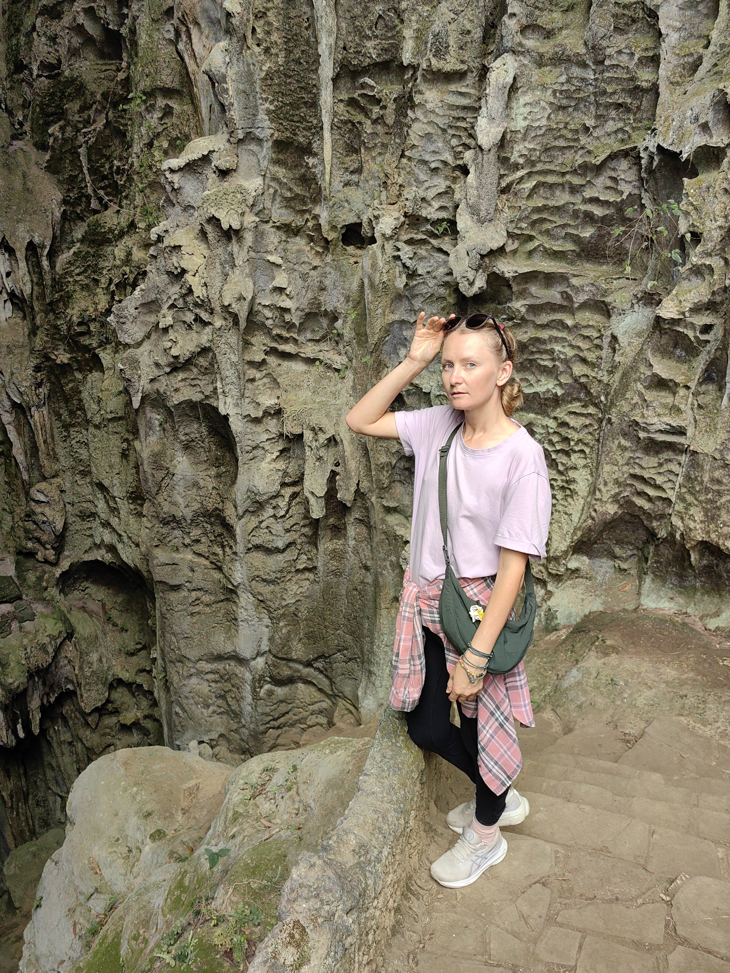 A woman stands on a stone staircase at Tam Thanh Cave, Lạng Sơn, Vietnam. She has fair skin, light hair styled in braids, and wears a lilac t-shirt, black leggings, and light-colored sneakers. A pink and white plaid shirt is tied around her waist, and she wears a green shoulder bag. Her left hand is raised to hold sunglasses on her head.
The background consists of a tall, textured rock face. A dark opening in the rock to the left suggests a cave entrance. The staircase is also made of textured stone, and some moss and small plants grow on the surrounding rocks.