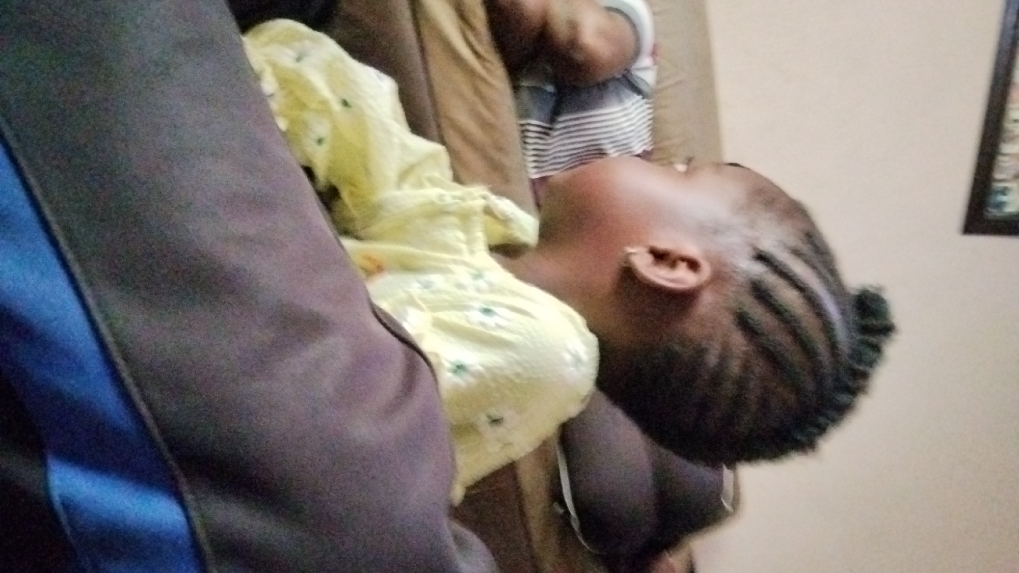 A child lies on a brown couch, with a person wearing a dark gray and blue jacket visible on the left side. The child has dark skin, braided hair, and wears a yellow floral top. Another person, wearing a striped shirt, is partially visible behind the child. A framed picture hangs on the wall in the background. The location is Jos, Nigeria.