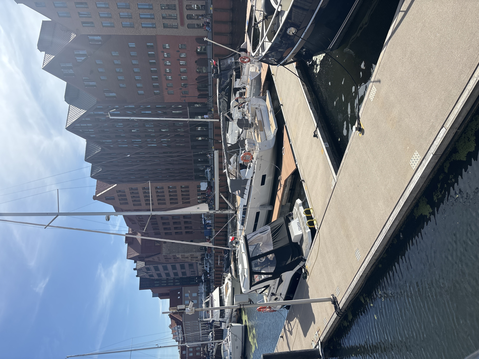 The image shows a marina with several sailboats docked. The sailboats are white with black accents and have their masts extended. One of the boats has a black cover over its cockpit. The marina is adjacent to a waterfront promenade made of concrete. In the background, there are multi-story brick buildings with many windows, some appearing to be residential or office spaces. The sky is blue with some wispy clouds, suggesting a clear day. The water in the marina is dark and reflects the sky and surrounding buildings, with some green algae visible on the surface. The scene appears to be calm and peaceful. The time of day seems to be daytime, likely late morning or early afternoon, given the bright sunlight casting shadows. Some text is visible on the back of one of the boats, reading "GDANSK".