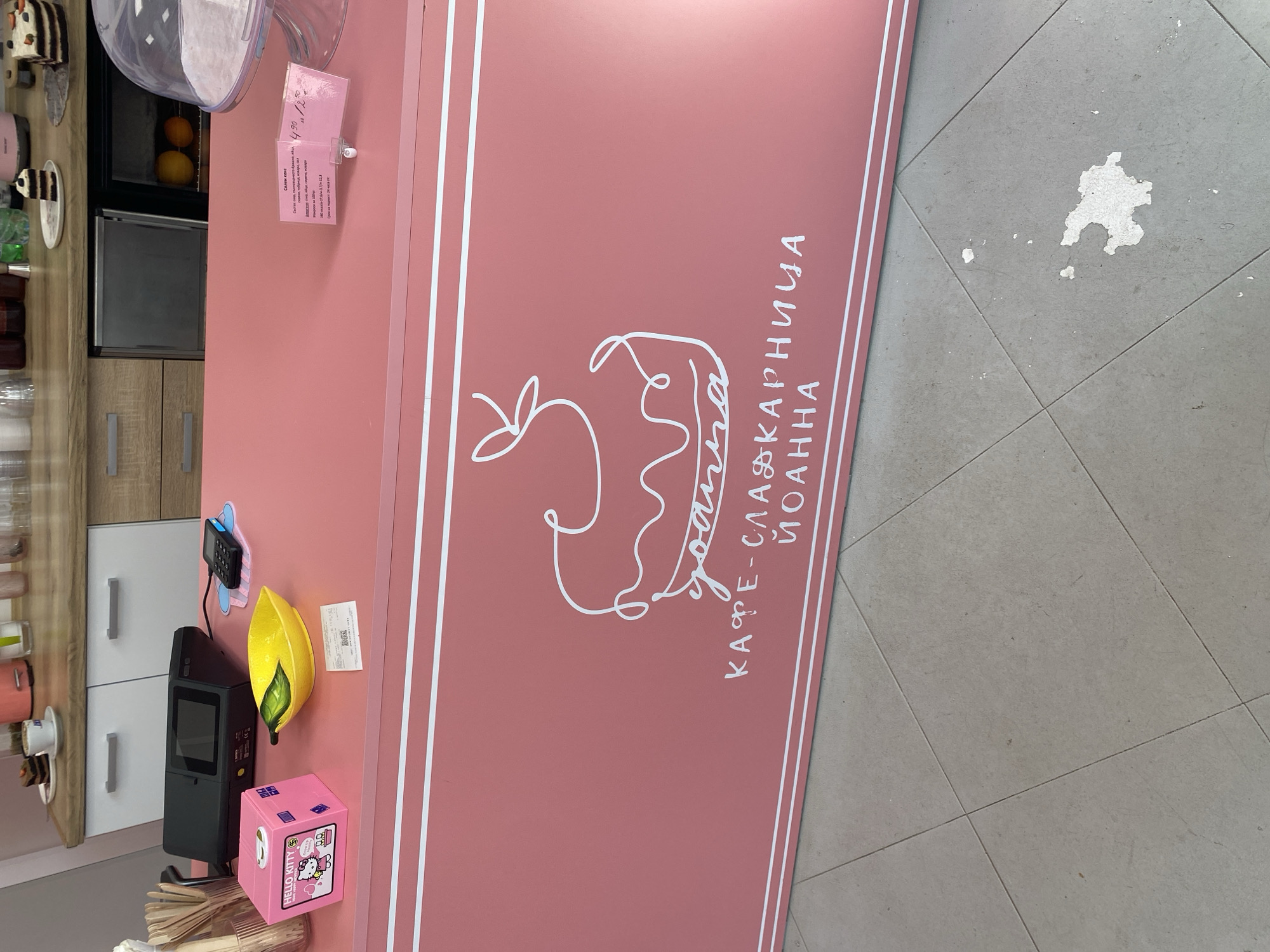 The image displays the interior counter area of a café-patisserie named "Йоанна" (Yoanna) located in Stara Zagora, Bulgaria.
The primary subject is a vibrant pink counter, the front of which features a white outline drawing of a dessert, incorporating the name "Йоанна" (Yoanna), with the full name "КАФЕ-СЛАДКАРНИЦА ЙОАННА" (Café-Patisserie Yoanna) written below in white Cyrillic capital letters. White double lines run along the top edge of the counter's front.
On the pink countertop, from left to right:
* A clear glass container holds numerous light-colored wooden stirring sticks.
* A pink square "Hello Kitty" branded box, labeled "HAPPY BIRTHDAY," is positioned next to the sticks.
* A yellow ceramic dish shaped like a lemon with a green leaf detail.
* A small white paper receipt lies flat on the counter.
* A black electronic point-of-sale (POS) terminal or cash register with a dark screen.
* A smaller black payment terminal or card reader rests on a light blue, cloud-shaped mat.
* Towards the right, a clear, dome-shaped cake stand cover is visible.
* A small, upright pink plastic sign holder displays a menu or price list in Bulgarian, showing "4.90 / 8.50" (likely prices).
Behind the counter, a light wood-toned surface serves as a backdrop, with white cabinets featuring silver horizontal bar handles beneath it. On the wooden surface:
* Various clear plastic cups and bottles with colored liquids are present.
* A white cup with a saucer and a slice of chocolate cake topped with a cherry.
* Another slice of multi-layered cake, possibly with nuts or fruit, is visible further right.
* A small under-counter refrigerator unit contains two oranges.
* Part of a pink kitchen appliance, possibly a blender or coffee machine, is visible on the far left.
The floor in the foreground consists of light grey square tiles with visible grout lines forming a grid pattern. Irregular white marks or debris are present on the tiles in the lower right portion of the image. No people are visible. The lighting is bright and even.