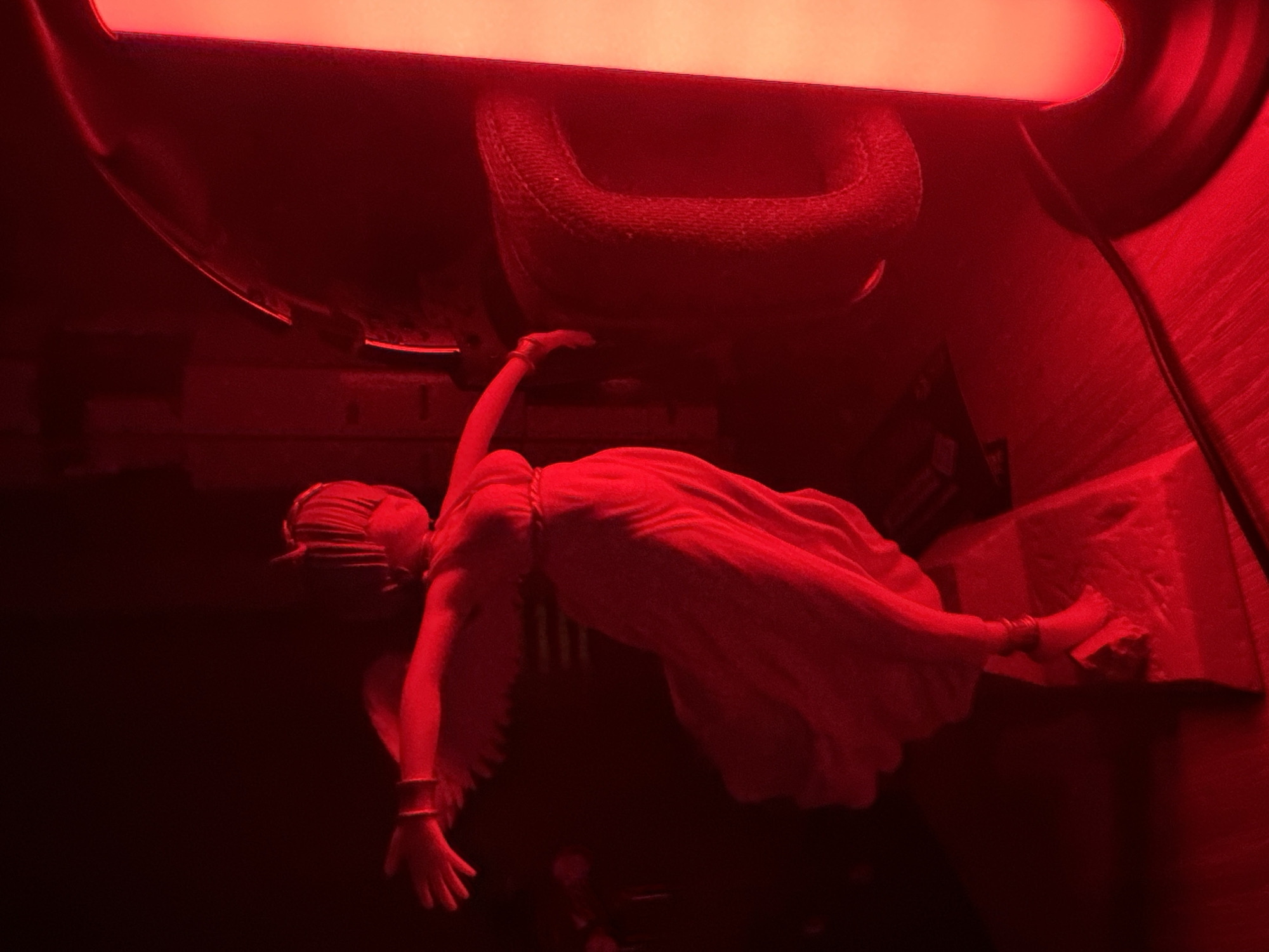 In a dimly lit environment, illuminated by a bright red light source on the right, a figurine of a winged female stands. The figurine is clad in a flowing, draped garment. Its arms are outstretched to the sides, and its head is tilted slightly back. The figurine is positioned on a raised platform. To the right of the figurine, a large, dark object with a textured surface, possibly headphones, is visible. A thin, black cable runs across the foreground, leading towards the red light source. The background is dark and indistinct, with some blurred shapes suggesting a room or indoor setting.