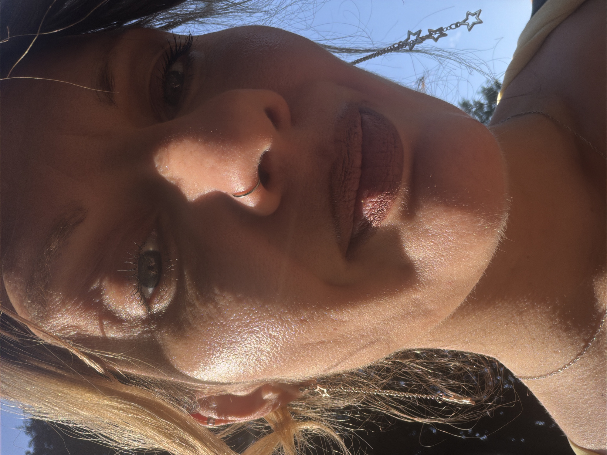 A close-up, low-angle shot focuses on a person's face, predominantly their right eye, nose with a hoop piercing, and lips. The individual has fair skin with visible pores and fine lines. Light reflects off their skin, creating highlights and shadows. Their right eye is partially obscured by a shadow, while the left eye is more visible. They have dark eyelashes and eyebrows. The lips appear to have a textured surface, possibly due to lipstick or lip gloss. Strands of light-colored hair frame the face, and a star-shaped earring dangles near the ear. Above, a blue sky is visible, and a necklace with star charms is partially visible. The background is blurred and indistinct, with hints of greenery and what appears to be the edge of clothing. No specific details indicating a location in London, United Kingdom, are discernible.