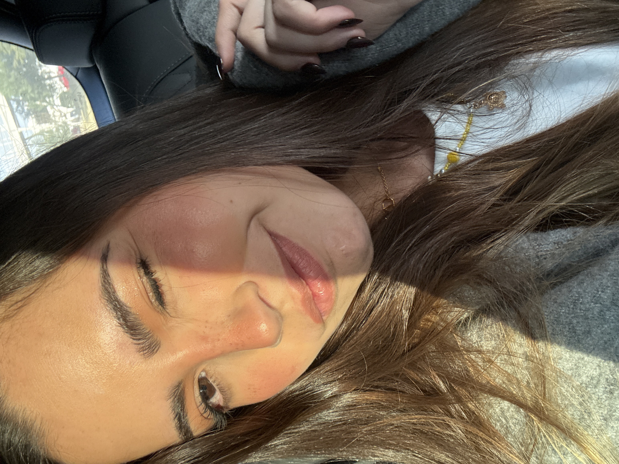A young woman with long brown hair is pictured indoors, seemingly in a vehicle given the view of the outside through the window behind her. She's wearing a light grey sweater and a white garment beneath. She has a golden necklace with a small charm. The lighting is bright, with a shadow partially obscuring her face. Her fingernails are painted in a dark color. No specific details in the image indicate a location in San Bernardino Tlaxcalancingo, Mexico.