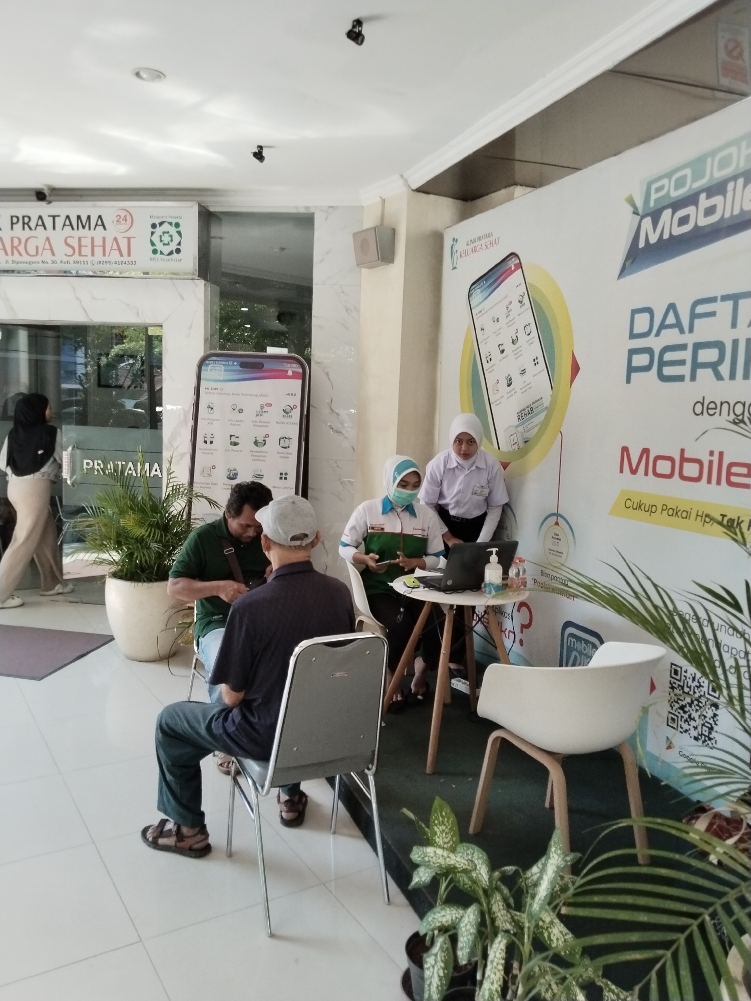 The image depicts the interior of a clinic, likely "Klinik Pratama Keluarga Sehat," located in Kecamatan Pati, Indonesia, as indicated by the signage. The scene features a registration or information booth with two women in medical attire attending to a visitor. One woman, wearing a white lab coat and hijab, is behind a small table with a laptop, hand sanitizer, and water bottles, appearing to be assisting the visitor. The other woman, also in medical attire and hijab, is seated and looking at a mobile phone.
In the foreground, two men are seated, facing away from the camera. One man, wearing a dark shirt and a grey cap, is seated in a grey chair. The other man, wearing a dark green shirt and a brown strap across his chest, is also seated, possibly in conversation with the man in the grey cap. Their posture suggests they are waiting.
A large advertisement or display board is prominently featured on the right side of the image, showcasing a smartphone interface with text in Indonesian, including "POJOK Mobile" and "DAFTAR PERIKSA." A giant replica of a smartphone is also displayed near the entrance. Plants are placed around the waiting area, adding to the environment. The overall setting appears to be a clean, well-lit indoor space, possibly a waiting area or a service point within the clinic.