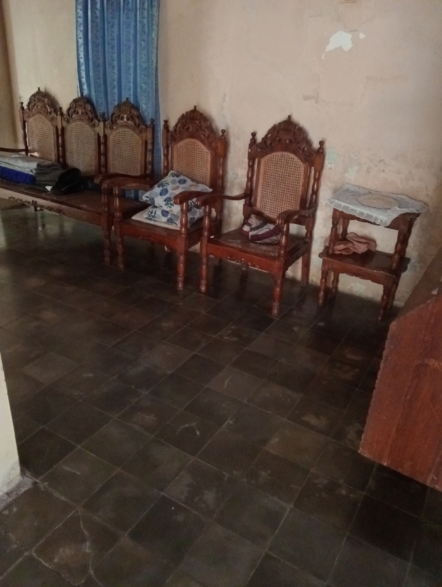 A dimly lit, indoor scene depicts a row of four ornate wooden chairs with woven cane backs, positioned against a textured, pale wall. To the left of the chairs, a long wooden bench or table is visible, draped with folded blankets and what appears to be a black hat. The chairs are separated by the typical gaps between furniture pieces, and the closest chair on the right has two cushions with a blue floral pattern. To the right of this chair, a small wooden side table with two shelves stands, holding a white, lacy cloth on its top surface and a bundle of fabric on the lower shelf. The floor is covered in dark, square tiles. A blue curtain is visible behind the chairs. The overall impression is of a simple, perhaps older, domestic interior. The location is identified as Krajan Kidul, Indonesia.