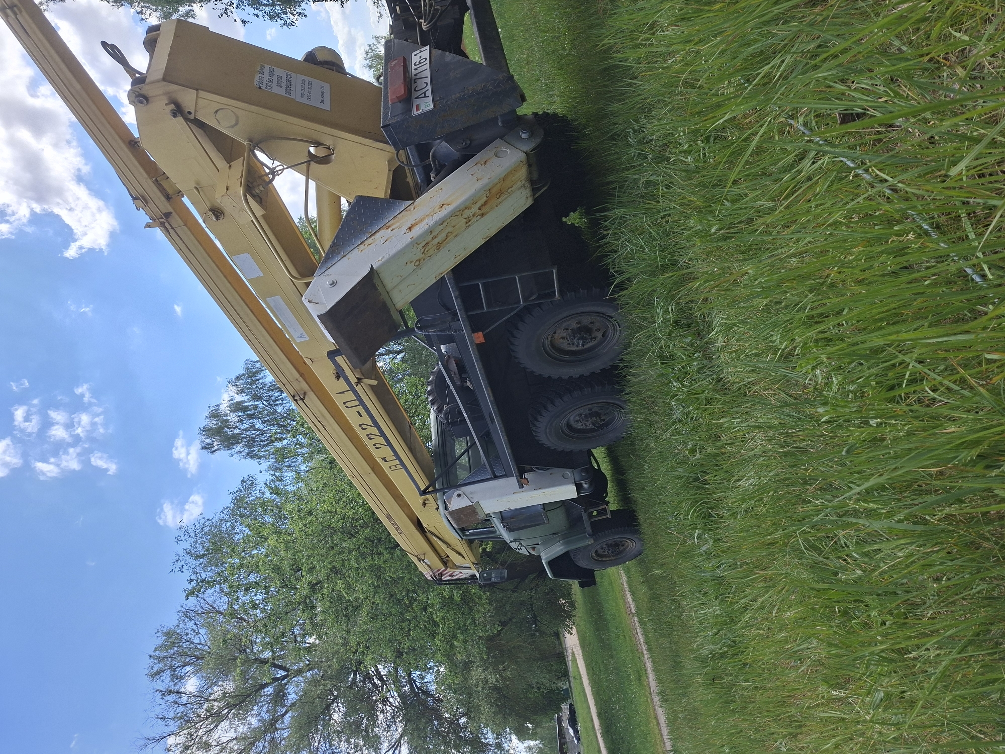 The image shows a yellow truck crane parked on grass, possibly in Zhabinka, Belarus, as indicated by the Belarusian license plate "AC7116-1". The boom is extended towards the sky, partially obscuring the view of trees and the partly cloudy sky. The truck has multiple wheels and appears to be heavy-duty. Text on the boom reads "БF 222-01" and provides safety instructions.