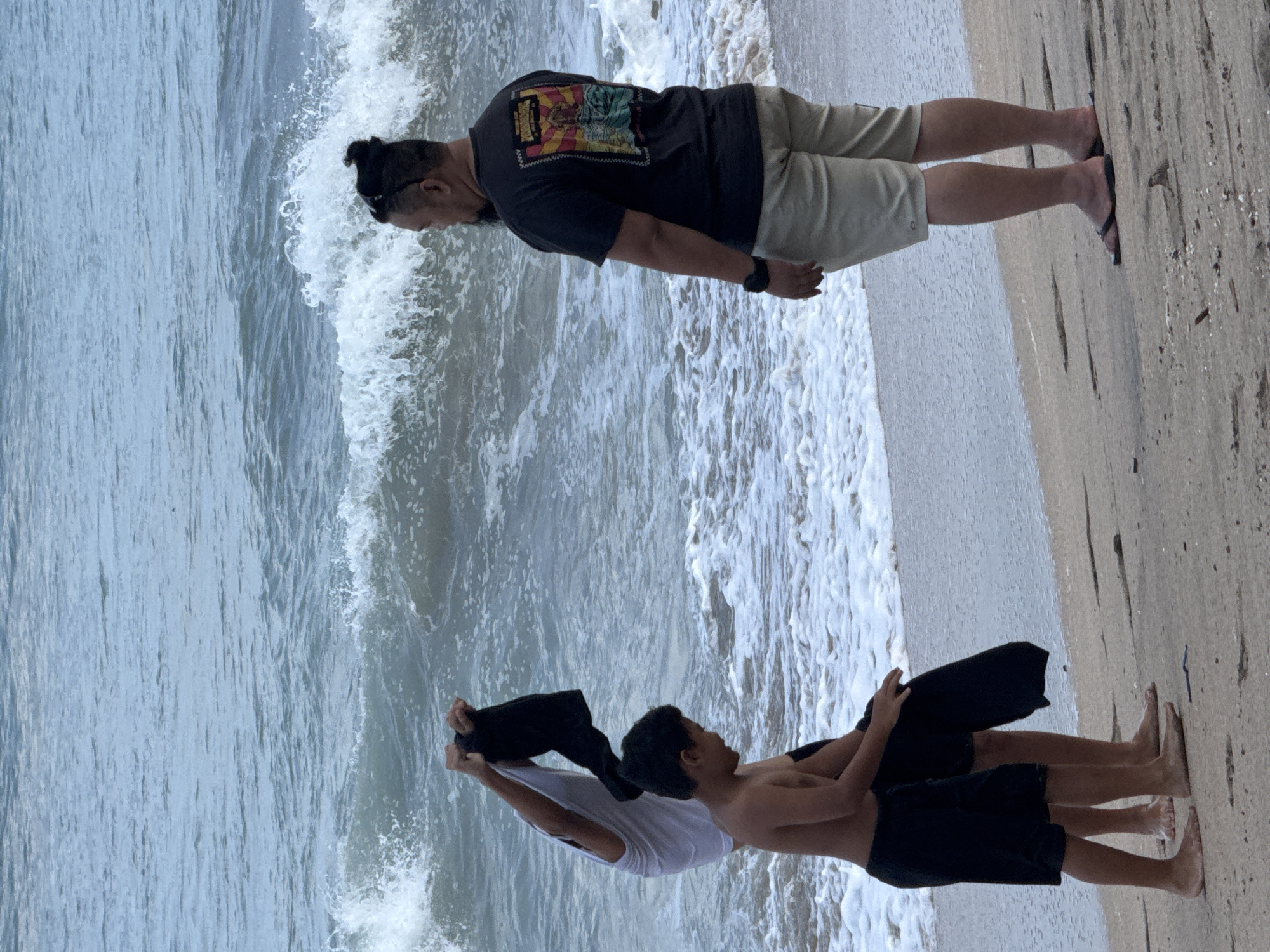 A medium shot captures three individuals on a sandy beach in Kuta, Indonesia, with ocean waves breaking in the background. On the left, a person wearing a white t-shirt is holding up a black garment, their arm extended. Beside them, a shirtless boy in black shorts is partially visible, facing away from the viewer. To the right, a man with a man bun, wearing a dark t-shirt with a colorful graphic and tan shorts, is seen from the back, looking out at the ocean. The waves are moderate, creating white foam along the shoreline. The sand is a light brown, with some debris scattered on its surface. The overall lighting suggests an overcast day.