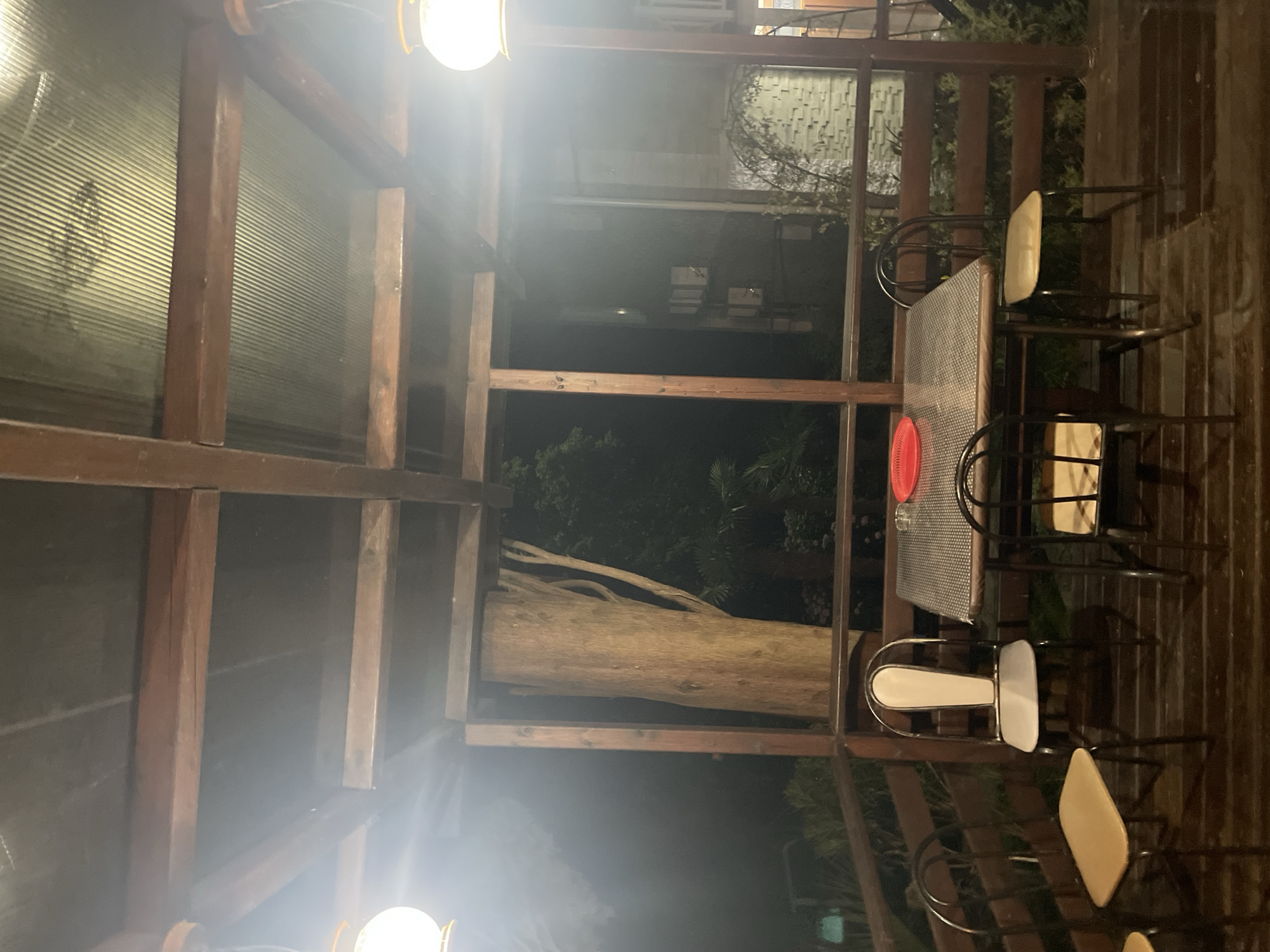 The image depicts an outdoor seating area at night, likely a covered patio or balcony. A square table with a patterned surface is set in the center, with four chairs around it. One chair has a white back and seat, while the other three have tan seats and dark frames. A red plate sits on the table. Wooden railings and supports frame the scene, and a large tree trunk is visible to the left. Beyond the immediate area, darkness prevails, with faint lights suggesting other structures or a distant view. Hanging lamps are visible in the upper corners, casting a warm glow. The ground is made of wooden planks.