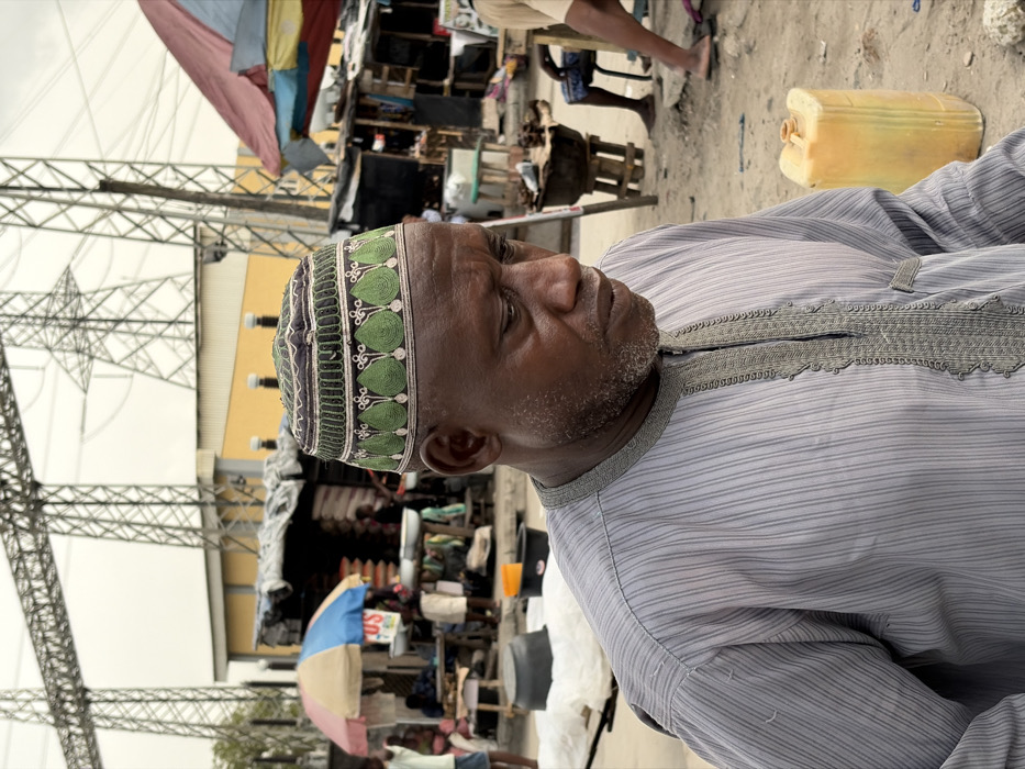 An adult Black male is depicted in a semi-close-up, looking downwards and to his right, in an outdoor market setting in Jakande, Nigeria. He wears a gray, vertically striped tunic with intricate gray embroidery around the placket and neckline, and a green and white embroidered kufi cap featuring leaf-like patterns. His short beard is dark with some gray.
Behind him, an active market scene unfolds under an overcast sky. To the left, a stall with a striped blue, white, and tan umbrella features white basins and stacked bags, with multiple individuals partially visible. To the right, another stall with a reddish tarp roof is present, with a person seated and a dark pot on a wooden stand. A yellow plastic jerrycan rests on the dusty, unpaved ground in the mid-right. In the far background, two large metal lattice electrical transmission towers rise above a yellow-walled building with a gray roof, indicating a proximity to power infrastructure.