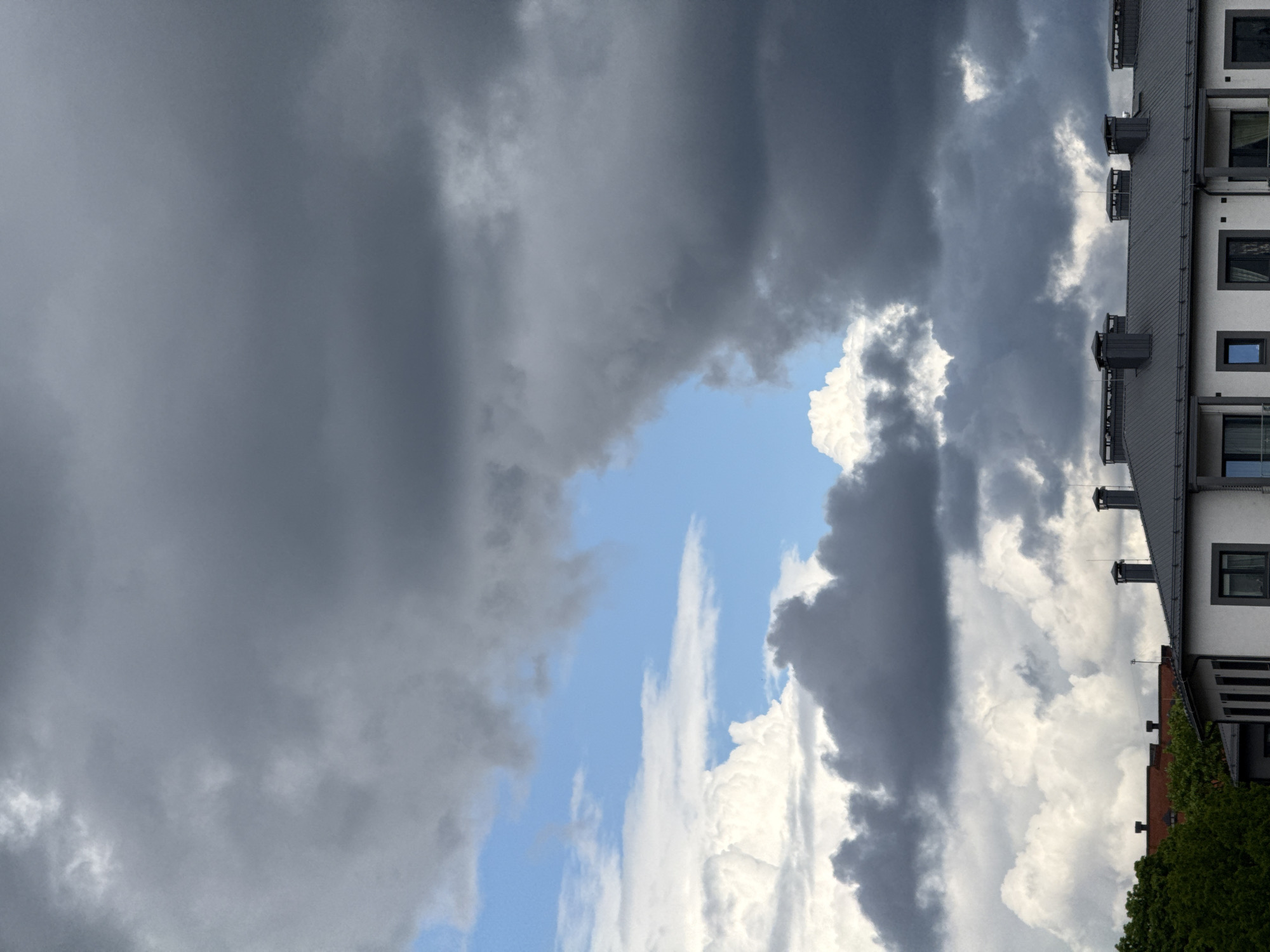 The image captures a view of Riga, Latvia, showcasing a sky filled with a mix of dark, heavy storm clouds and patches of bright blue sky. A building, possibly an apartment complex, is partially visible on the right side of the frame, with its roofline, windows, and a glimpse of greenery at its base. The lighting suggests a dynamic weather situation, with the sun intermittently breaking through the cloud cover. There are no people visible in the image. The scene conveys a sense of impending rain or a recent storm.