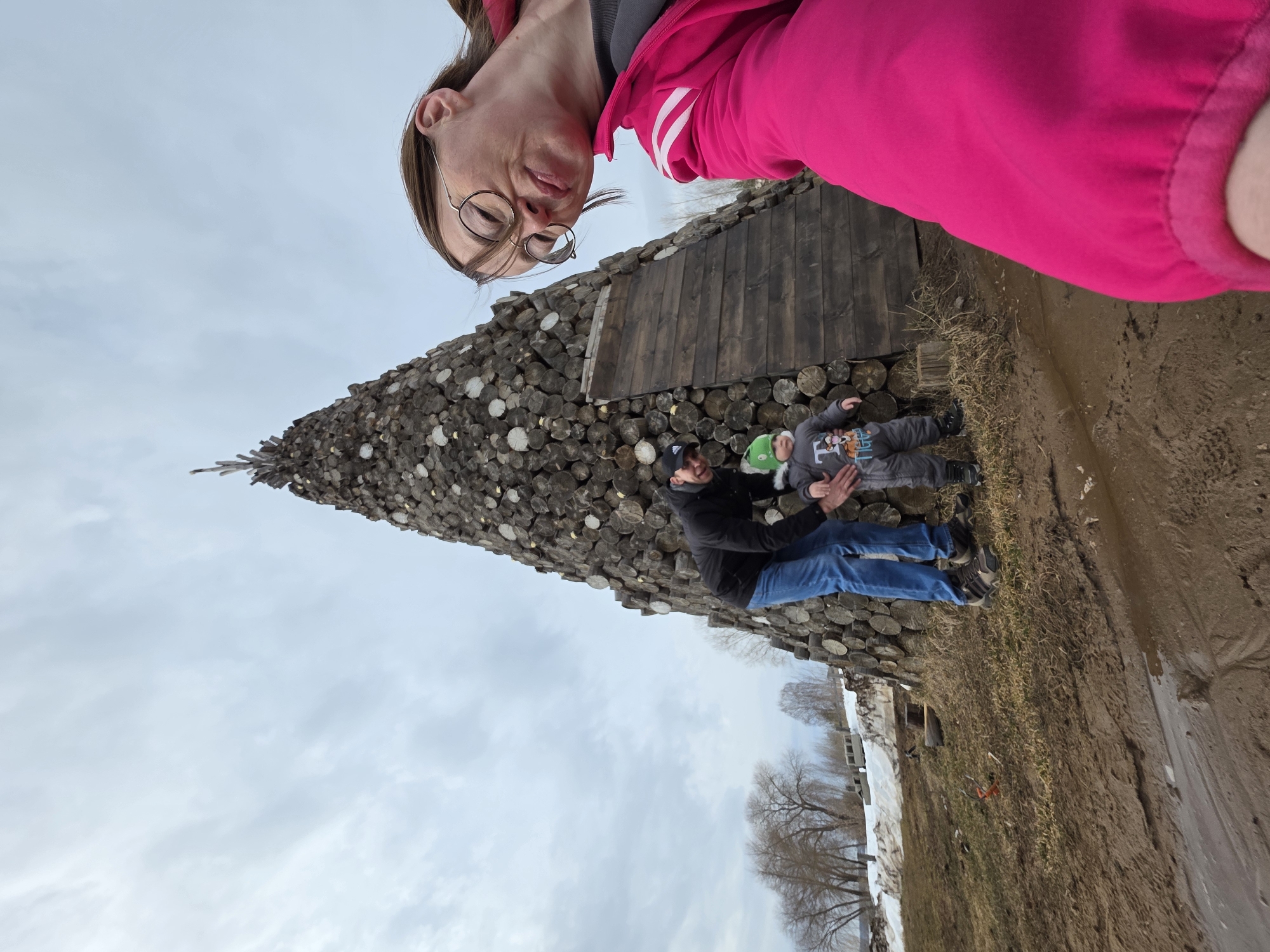 A medium shot, taken at an unusual angle, depicts three people and a conical structure in an outdoor environment in Votkinsk, Russia. A woman with glasses, wearing a bright pink jacket, is positioned in the upper right of the frame. Below her, against the backdrop of a conical stack of firewood, a man is interacting with a child lying on the ground. The conical structure, made from stacked logs, tapers to a point, giving it a tree-like shape. The ground is a mix of dirt, grass, and some snow. Bare trees and a cloudy sky complete the background. The composition appears as if the camera was rotated counterclockwise, giving an upside-down orientation.