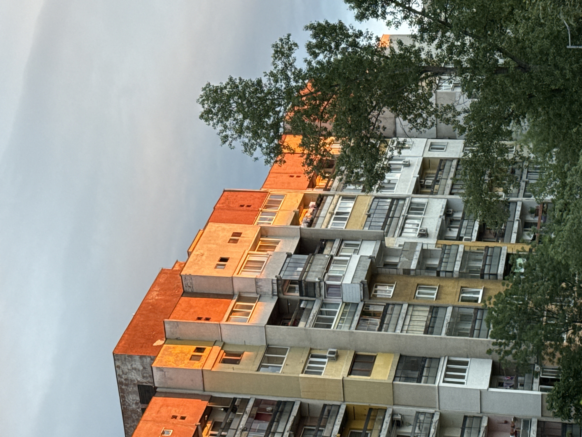 A low-angle shot depicts a multi-story building in Sofia, Bulgaria, likely an apartment complex, seen under an overcast sky. The building's facade displays a block-like design, with varied pastel hues and shades of gray and beige, with accents of red on the upper levels. Numerous windows of different styles are visible, and some units have air conditioning units attached. Parts of the building appear to be illuminated by a golden light, possibly from the setting sun. The foliage of trees partially obscures the building on the right side and in the top center.