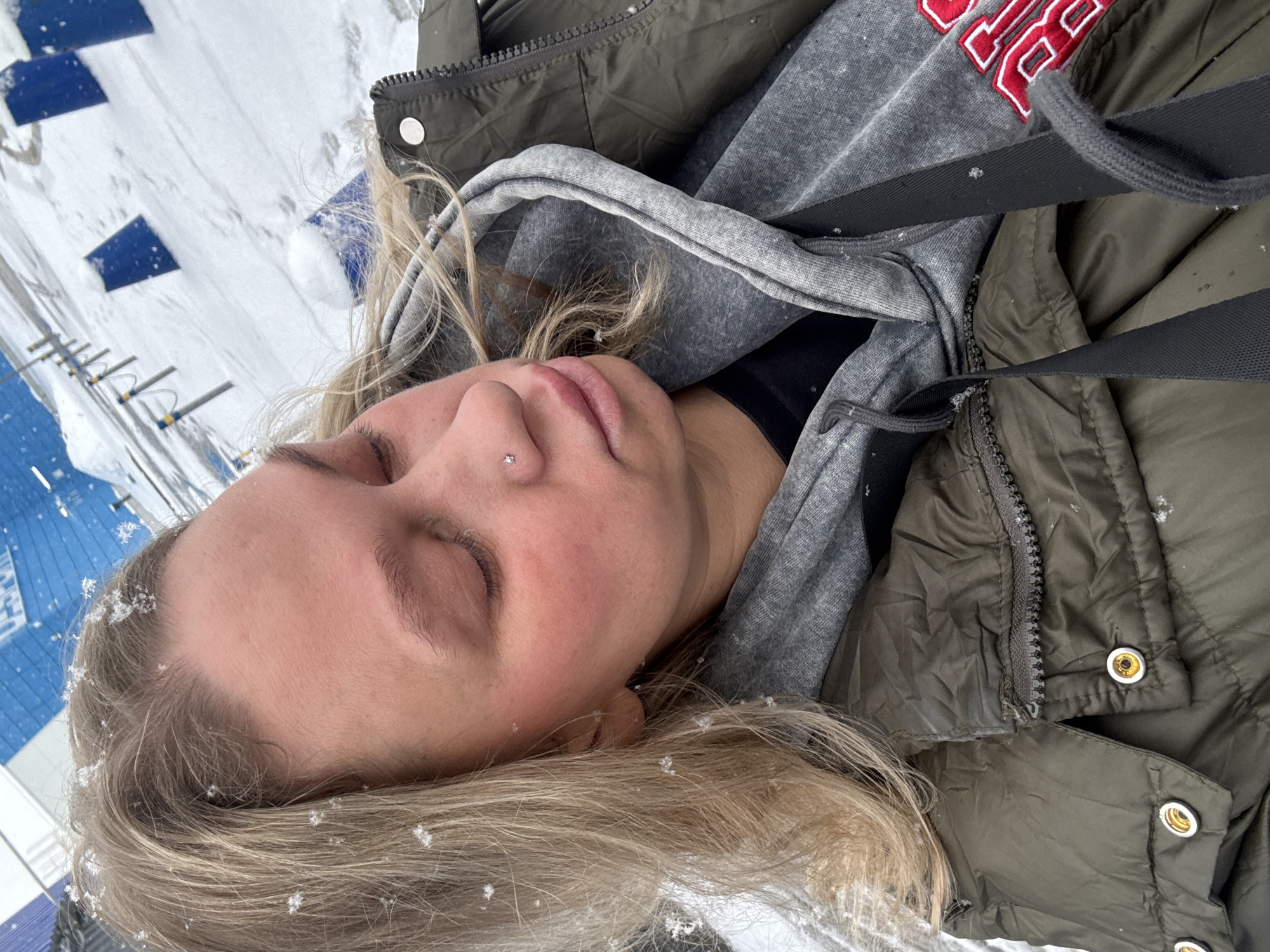 A young fair-skinned individual with blonde hair is lying on a snow-covered surface, with snowflakes visible on their hair and face. They are wearing a gray hoodie with red lettering partially visible, a black top underneath, and an olive-green puffer jacket. A black strap is crossing their body. The background shows snow-covered ground, a blue and white structure, and metal posts, suggesting an outdoor environment possibly in the city of Perm, Russia. The person's eyes are closed.