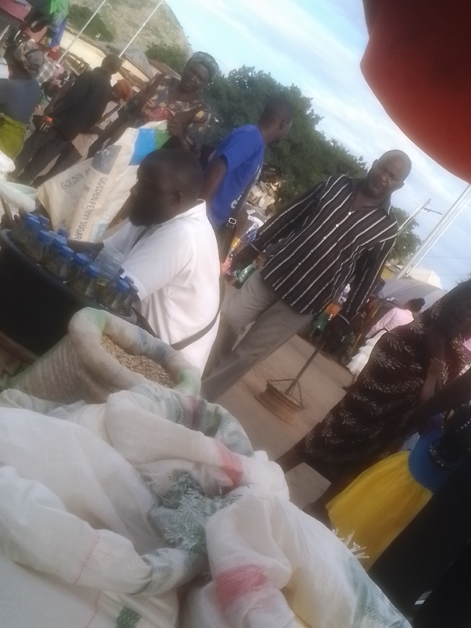 The image depicts an outdoor market scene, possibly in Gwong, Nigeria, with a tilted perspective. Several individuals are present, engaged in various activities. In the foreground, a man in a white shirt with a black strap is partially visible, and a large sack filled with granular material, possibly beans or seeds, is in front of him. To his left, another sack, also containing granular material, is visible. Further back, a man in a blue shirt is visible, and a man in a horizontally striped shirt and light-colored pants is also present. A woman wearing a patterned dress is visible on the right side of the image, and another woman in the background appears to be holding a child. Several bags, including one with "GOLDEN PENNY SUGAR" printed on it, are scattered around. The environment suggests a busy marketplace with trees and a cloudy sky in the background.