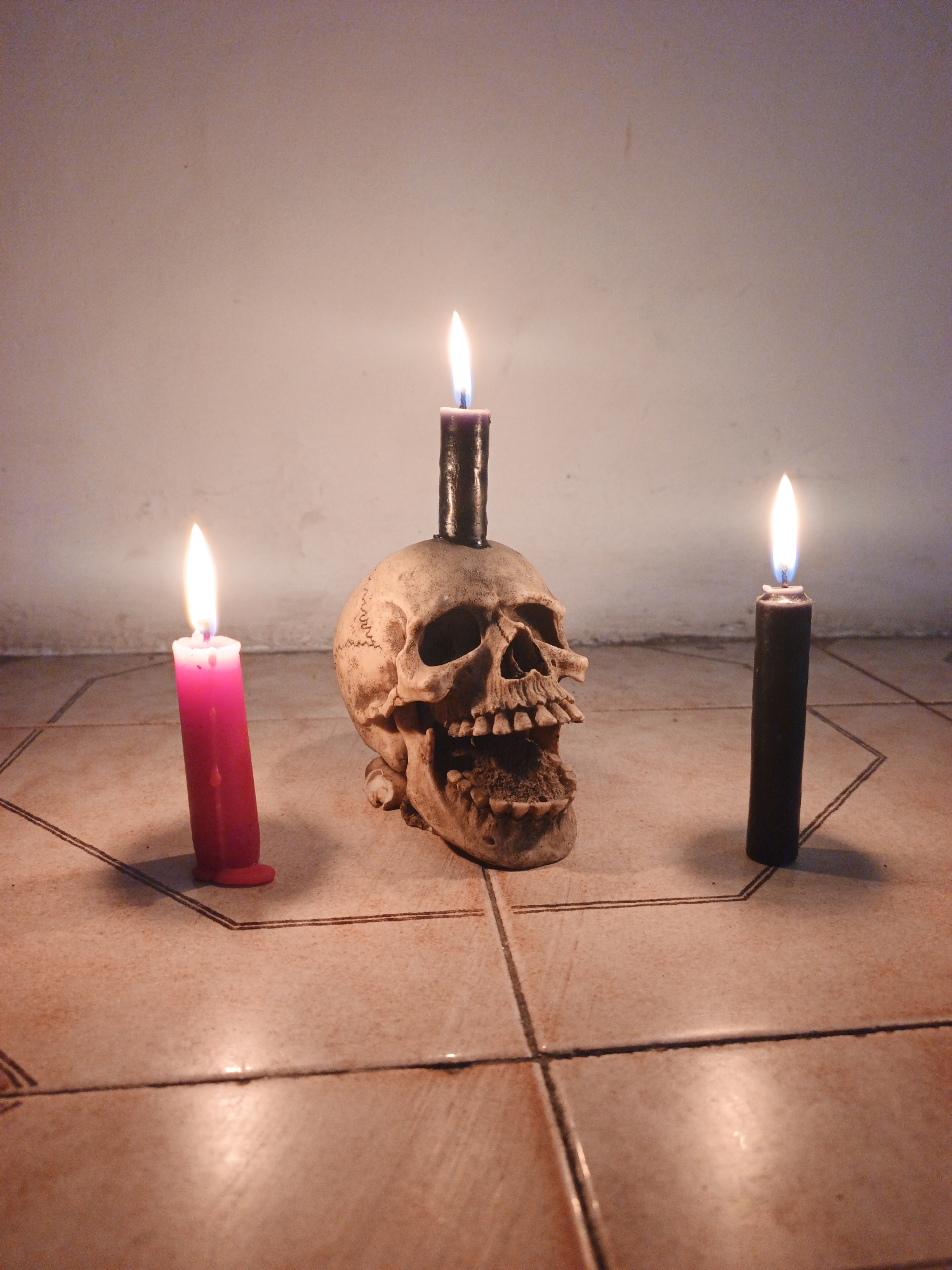 The image depicts a skull with a black candle stuck in its top. A pink candle is to the left, and another black candle is to the right, all three candles are lit. The scene is set on a tiled floor, with what appears to be a partial pentagram drawn around the items. The background is a plain wall. The location, Tinaquillo, Venezuela, is stated but is not verifiable from the image.