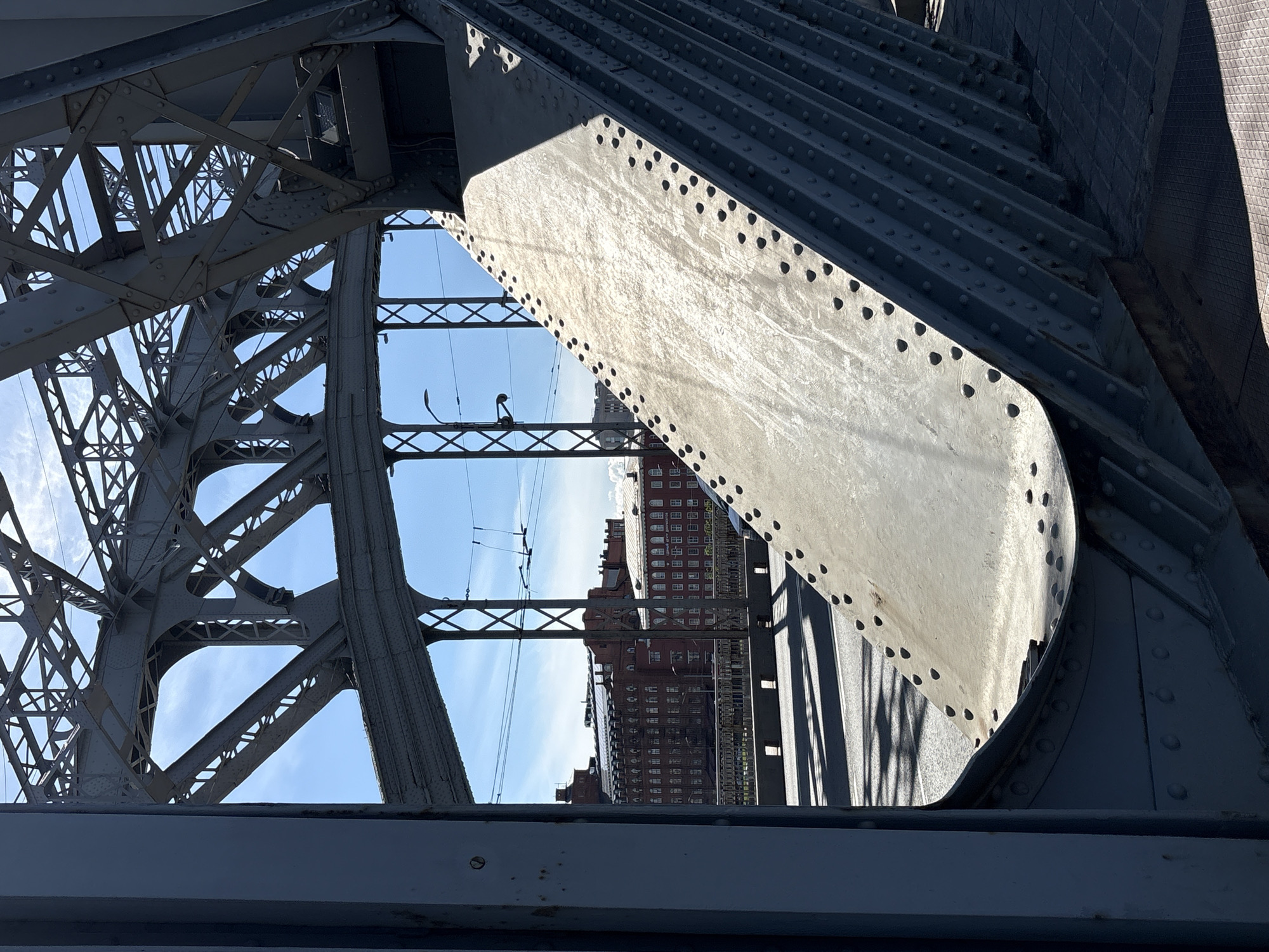This is a medium shot, eye-level photograph taken from under a large, riveted metal bridge structure. The bridge's intricate, dark gray metalwork dominates the foreground and mid-ground, creating a geometric framework through which the scene beyond is viewed. The sun is bright, casting strong shadows from the bridge onto the road below, suggesting it's either morning or late afternoon. The sky is a clear, bright blue with a few wispy clouds, indicating fair weather.
Through the gaps in the bridge's structure, a street is visible with a road and what appears to be a sidewalk. Several red brick buildings with many windows are lined up along the street, characteristic of urban architecture in Saint Petersburg. The overall impression is a bustling city scene viewed from a unique perspective beneath an industrial monument. There is no text visible in the image. The scene conveys a sense of everyday urban life framed by the impressive scale and design of the bridge.