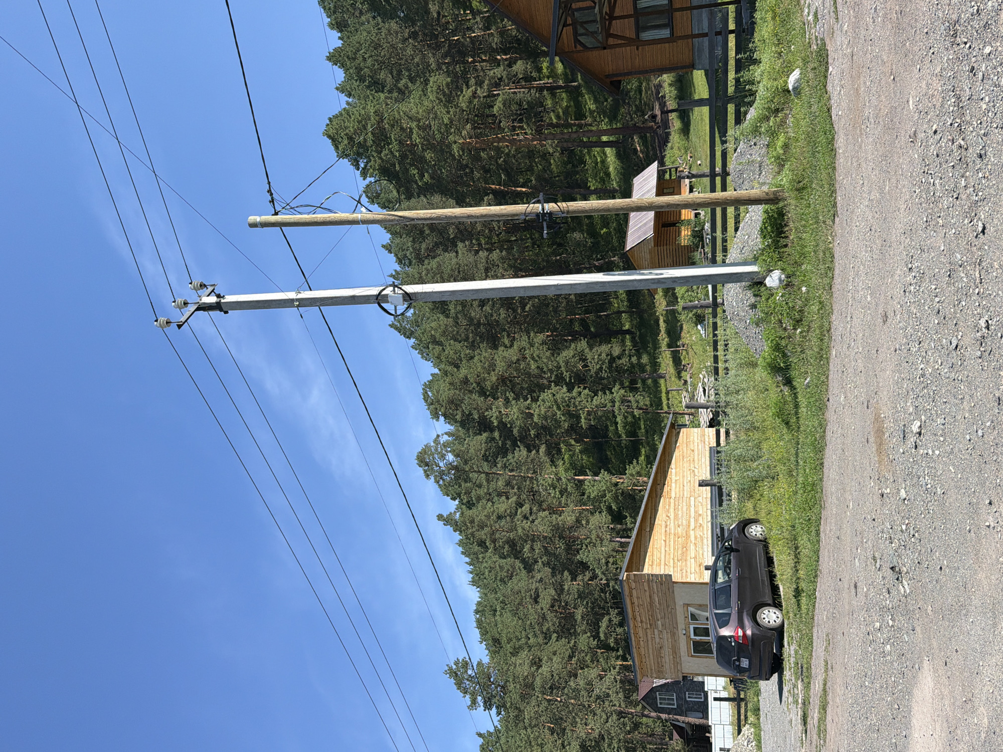 In Cheposh, Russia, a gravel driveway leads past a dark-colored hatchback car parked on grassy terrain. To the left, a modern wooden building with a beige base and a light brown wooden upper section stands under a clear blue sky. Behind the car and building, a dense forest of tall green pine trees covers a hill. Power lines stretch across the frame, supported by two utility poles: one concrete and one wooden. A small wooden structure with a metal roof is visible further up the hill, nestled among the trees, and part of a wooden fence can be seen.