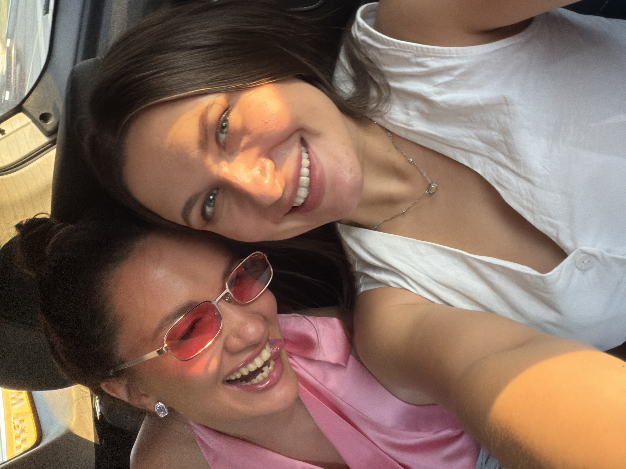 Two young women are in the back seat of a vehicle, taking a selfie during what appears to be sunset. The woman on the left is wearing pink clothing and pink-tinted sunglasses, with her hair in a bun. She is smiling and her tongue is slightly sticking out. The woman on the right is wearing a white top and a silver necklace with a small pendant. She is also smiling, with sunlight illuminating her face. The background shows the interior of a car, including a headrest and the rear window, with a blurred view of the outside. The overall lighting suggests it is late afternoon or early evening. The location is the city of Odintsovo, Russia.