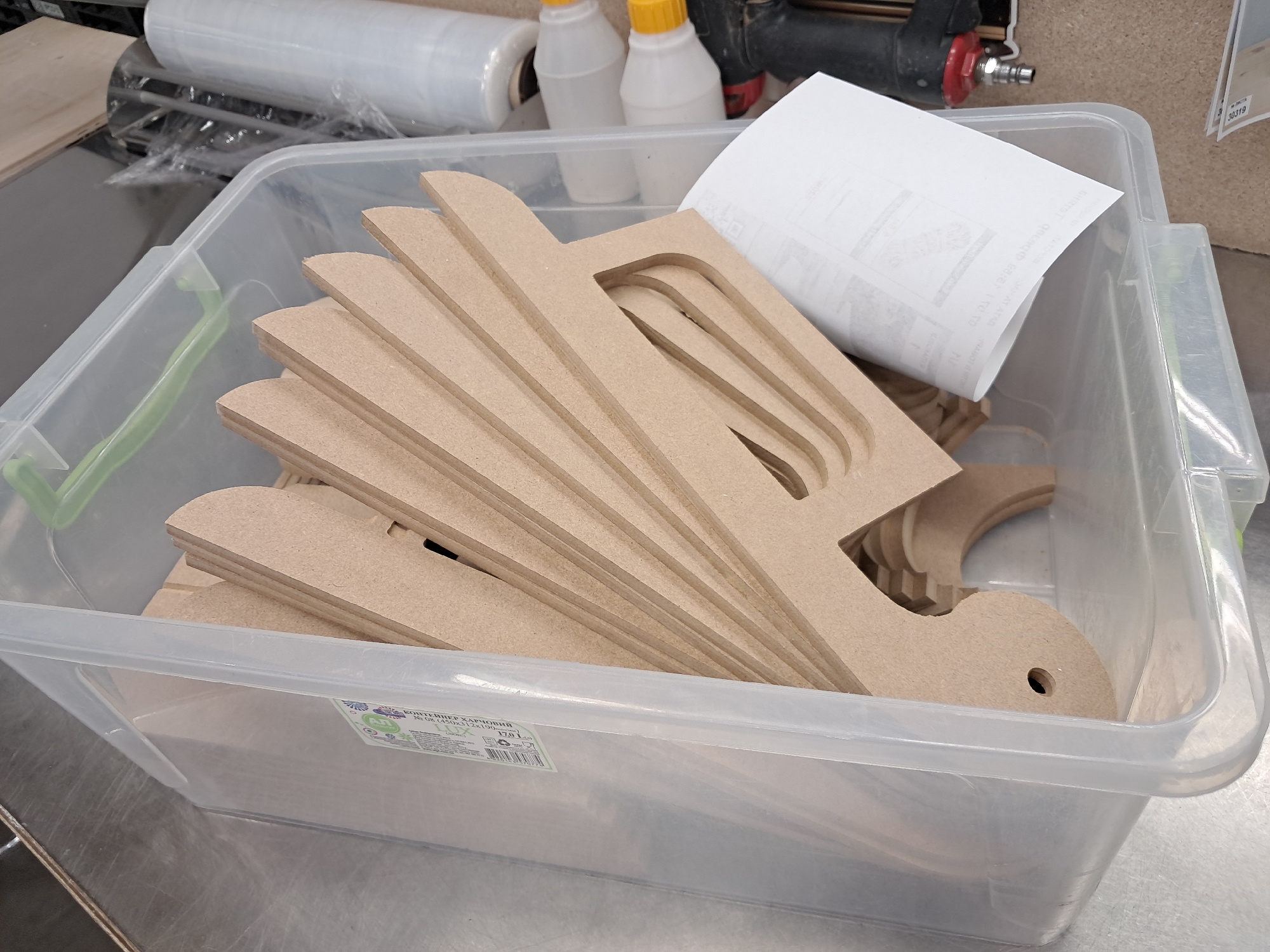A clear plastic storage bin is filled with stacked, wooden, laser-cut pieces. The pieces appear to be flat with a light brown, unfinished wood texture. Some pieces have cut-out shapes and a hole. A white piece of paper, possibly instructions or a label, is partially visible in the bin, folded over the wooden pieces. In the background, a roll of plastic wrap, two small plastic bottles with yellow caps, and what appears to be a pneumatic tool are visible. The storage bin sits on a metallic surface, likely a workbench. The scene is indoors.