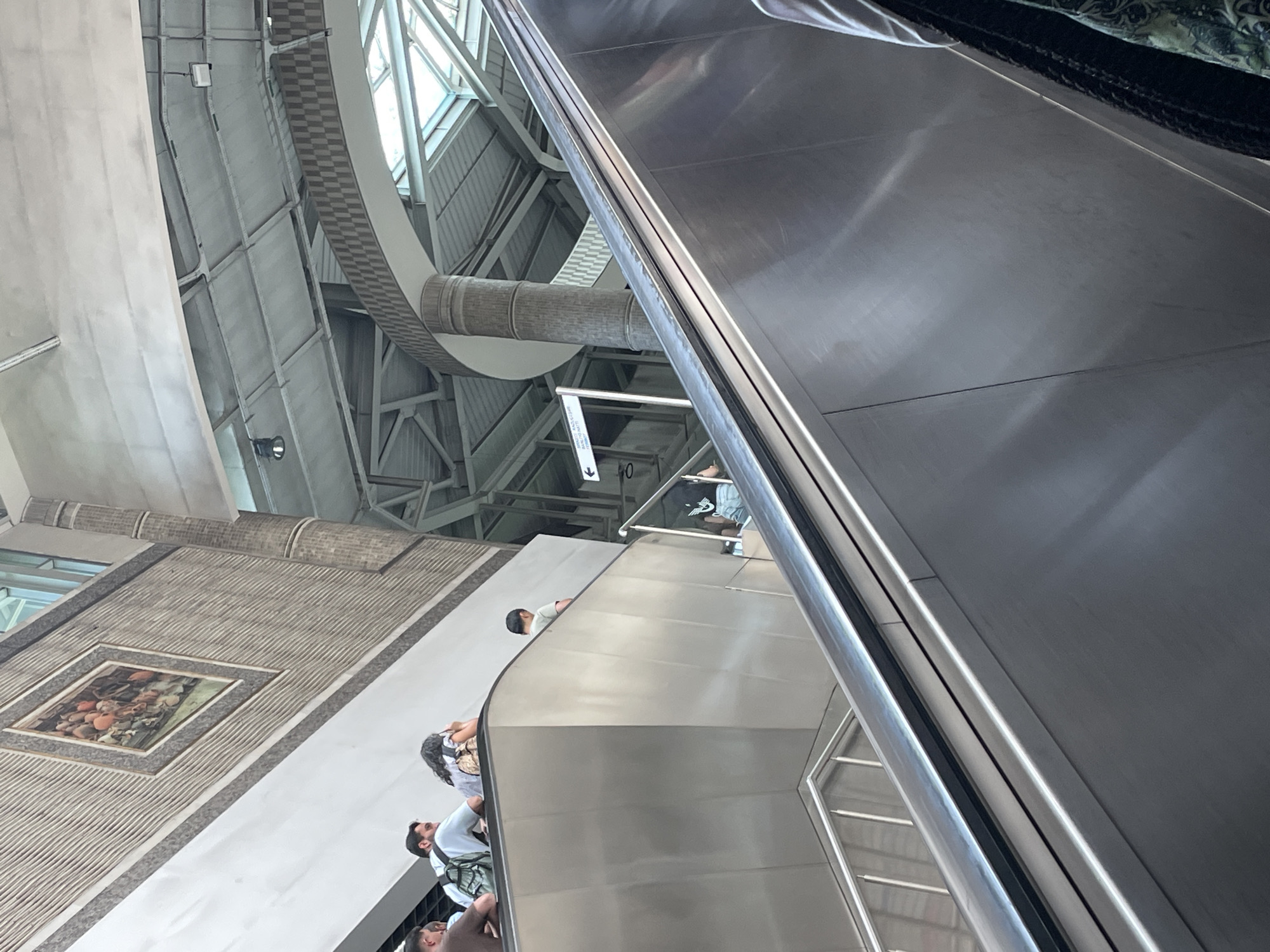 An upward-angled, medium shot captures people on an escalator within a modern, indoor transit hub, likely a train or metro station. The escalator's polished steel handrails and steps are prominent in the foreground. Above, the ceiling features exposed metal beams and a circular, checkered archway. To the left, a wall is adorned with a framed, rustic painting. Several individuals are visible on the escalator and the landing area. One person, facing away, wears a light blue shirt and a backpack. Another, with dark curly hair, is seen from behind. A child in a light green shirt stands on the escalator's upper level. A sign with an arrow points to the left, indicating directions within the facility. The overall environment suggests a bustling public space designed for efficient movement. The artwork on the wall and the architectural style are consistent with design elements found in Istanbul's newer transit infrastructure.