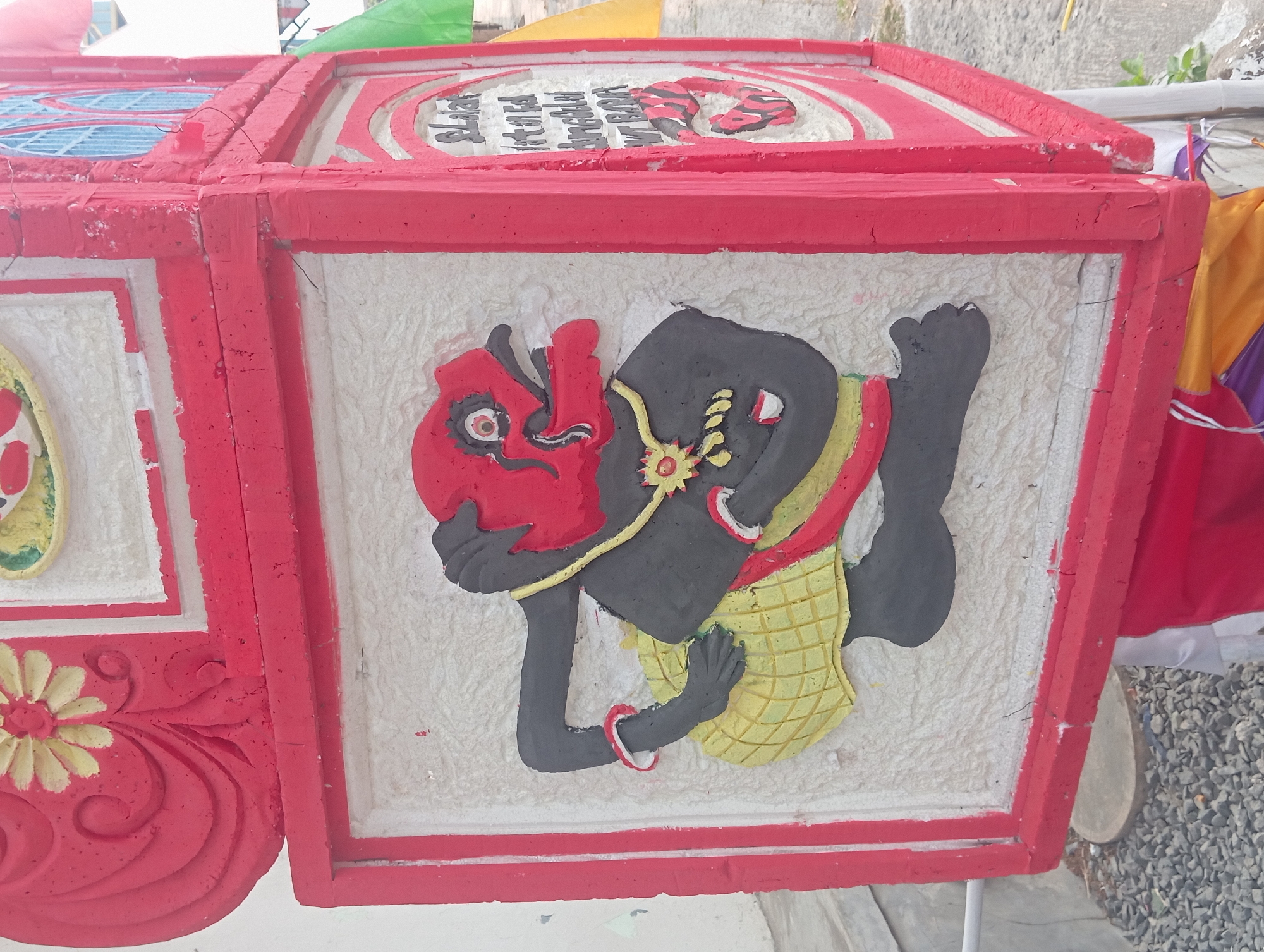 The image shows a brightly colored, three-dimensional artwork mounted on a pillar in Sokanegara, Indonesia. The artwork depicts a stylized figure with a red head and black body, wearing a yellow skirt and red armbands. The figure is in a dynamic pose, with one arm raised and the other bent at the elbow. The artwork is set against a white textured background within a red frame. To the right of this figure, another panel features text in white lettering on a red background, with some of the text appearing to be in Indonesian script. The overall structure is red and appears to be part of a larger decorative installation. The background shows a white wall, gravel, and some greenery, indicating an outdoor urban setting.