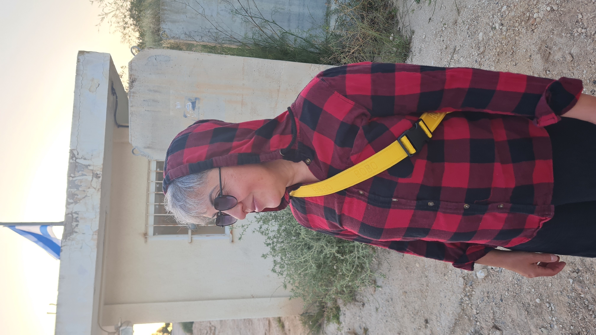 A medium shot captures a woman in a red and black checkered hooded shirt, accessorized with a bright yellow crossbody bag. She's wearing dark sunglasses. She is standing outside a building with a blue and white flag. The setting appears to be the city of Lachish Regional Council, Israel. The surrounding area features sparse vegetation and a sandy or gravelly ground.
