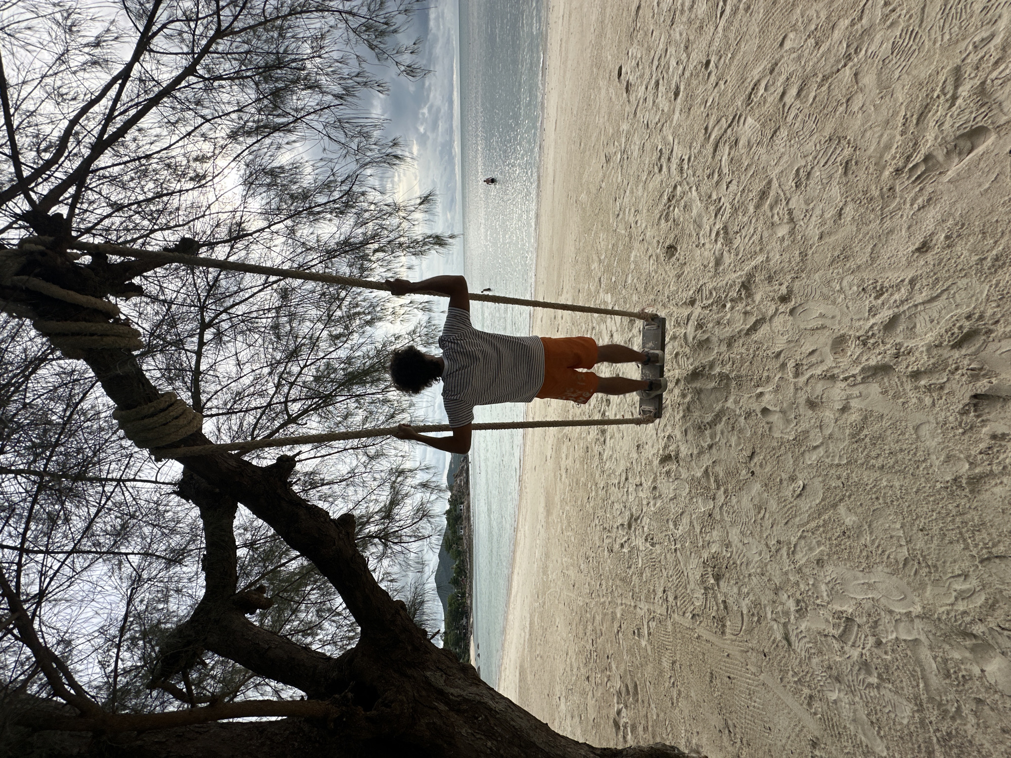 A person is seen from behind, sitting on a swing suspended from a tree by ropes. The person is wearing an orange and white striped shirt and orange shorts. They are facing the ocean, with a sandy beach in the foreground and lush green hills in the distance. The sky is overcast with dark clouds, and the sea is calm. The scene appears to be a peaceful, tropical setting on Ko Pha-ngan District, Thailand. There are two other people in the water, further out in the ocean. The sand is covered with footprints and tire tracks.