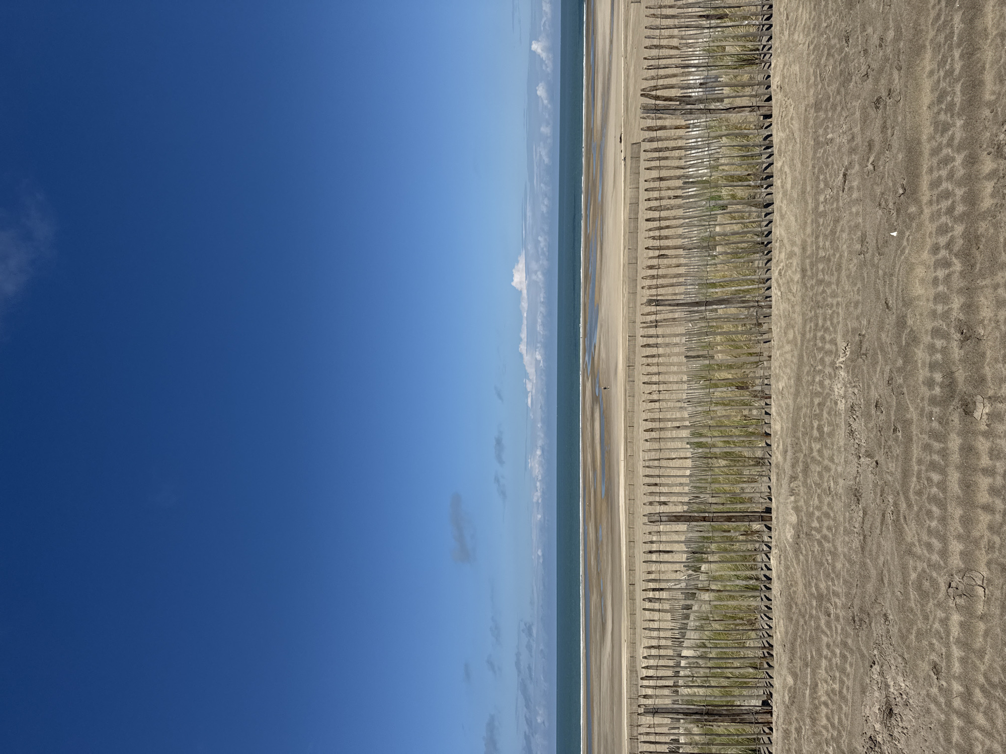 This image depicts a beach scene in Soulac-sur-Mer, France. The view is oriented with the sea and sky to the left, and the beach stretching to the right. The sky is a clear blue, with some wispy clouds near the horizon. The sea appears calm. Along the beach, a wooden fence runs parallel to the shoreline, with some vegetation growing between the fence posts. The sandy beach covers the rest of the image. The sand is textured with footprints and patterns. There are no people visible.