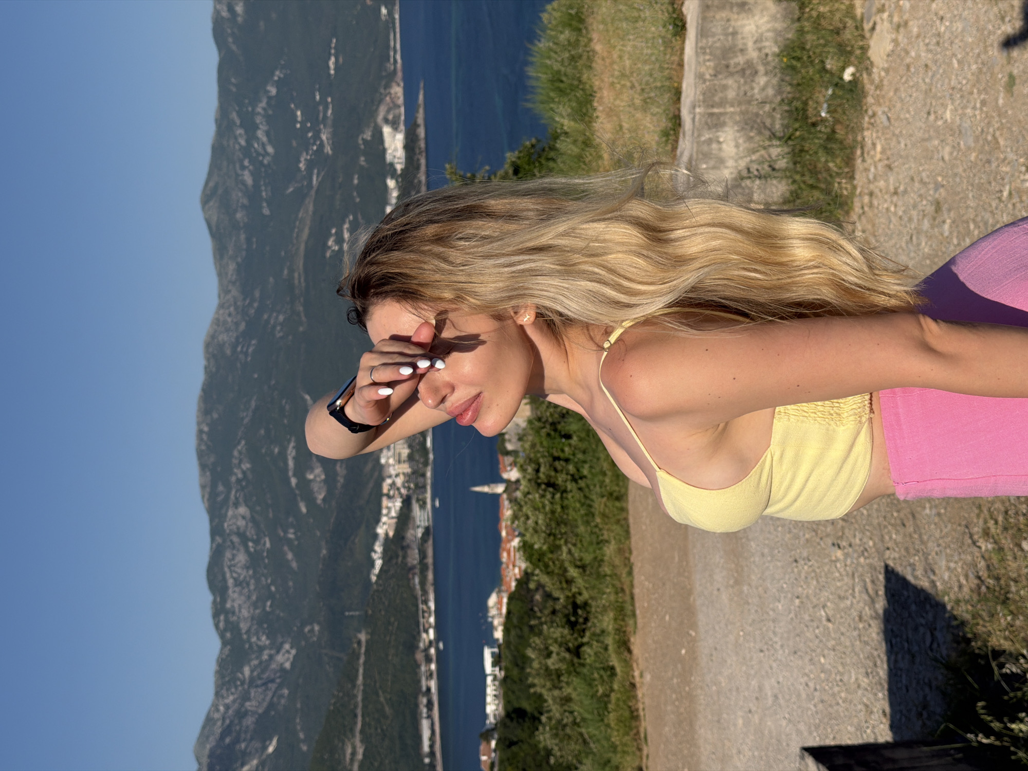 A woman with long blonde hair is standing on a stone path with green vegetation to her right. She is wearing a yellow top and pink pants and has a watch on her left wrist. She is shielding her eyes from the sun with her left hand.
Behind her is a scenic view of Budva, Montenegro. The blue sea and the rocky coastline, where buildings and vegetation are visible, stretch out in the background. A large mountain range rises to the left of the coastal area. The sky is clear and blue.