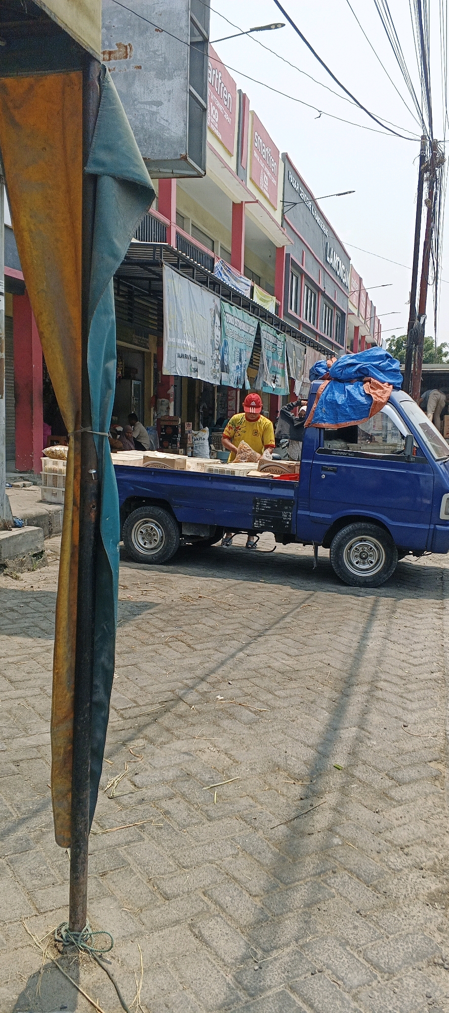 In Sumberwudi, Indonesia, a blue pickup truck is parked on a cobblestone street. A person wearing a yellow shirt and a red cap is standing in the back of the truck, sorting through a pile of items. The truck is loaded with several white crates and some boxes. In the background, a building with red trim and advertisements for "smartfren" is visible. A tall metal pole with a tattered fabric attached is in the foreground. The overall scene appears to be a market or a commercial area.