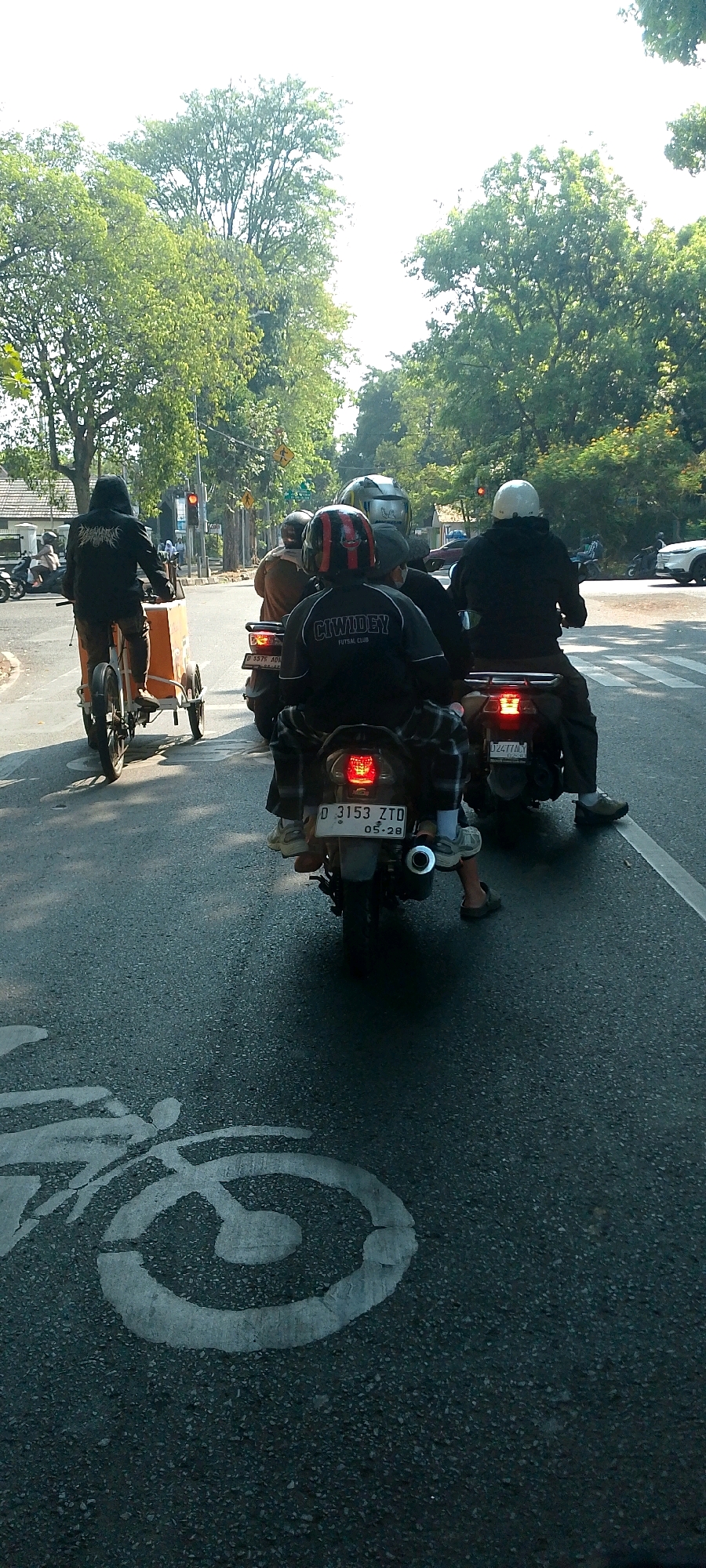 The image depicts a street scene in Bandung City, Indonesia, with a focus on several motorcycles and their riders on a paved road. In the foreground, a white bicycle lane symbol is painted on the asphalt. Several motorcycles are visible, some carrying multiple passengers. One motorcycle in the foreground has a license plate that reads "D 3153 ZTD 05-28". Another motorcycle behind it has a license plate that is partially visible. Trees line the street, and there are buildings in the background. The sky is bright, suggesting it is daytime. The scene appears to be at an intersection, with traffic lights visible in the distance. The overall impression is of everyday urban traffic in Indonesia.