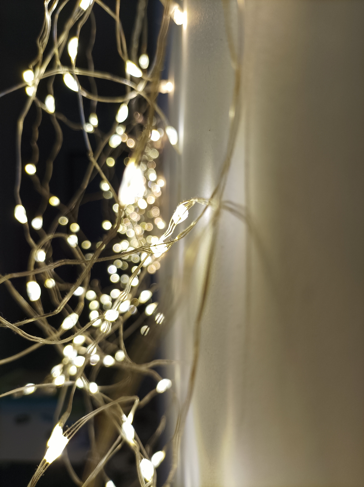 Close-up photograph of a string of small, illuminated LED lights clustered near a wall. The lights emit a warm, yellow-white glow and are attached to thin wires. The wall appears to be light-colored. The background fades into a dark space. The photograph does not show the specific location in Batumi, Georgia. The image provides no details about people, actions, or interactions.