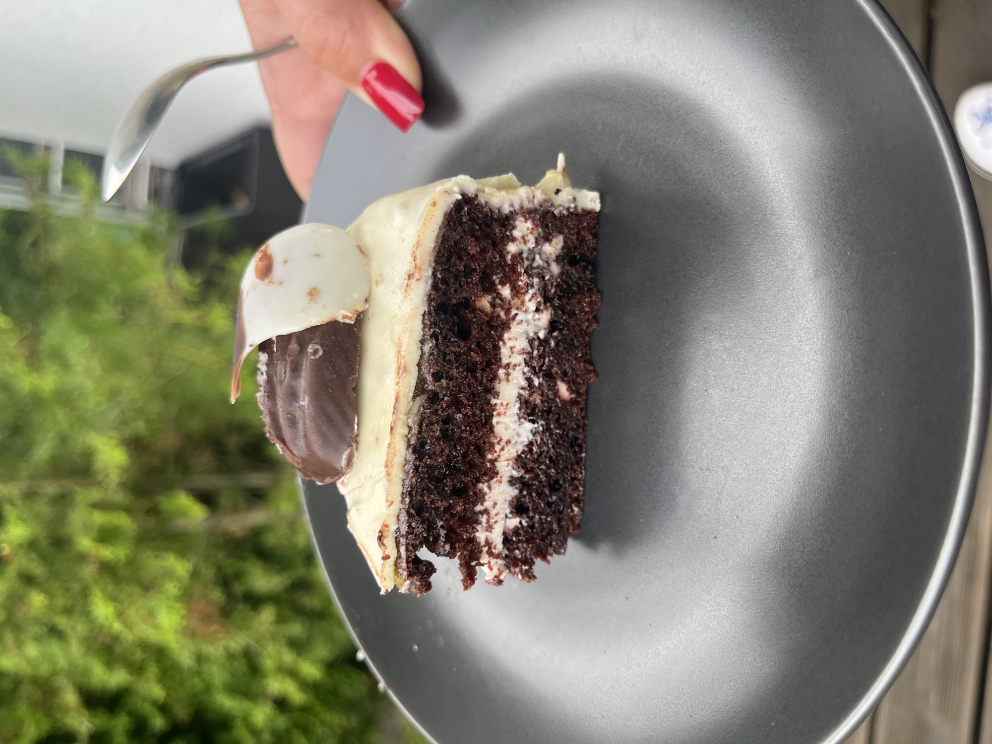 A slice of dark chocolate cake with white frosting and a dark chocolate decoration is presented on a black plate. The cake is layered with white cream. A person's hand, with bright red nail polish, is holding a silver spoon, poised to take a bite of the cake. The background features blurred greenery, suggesting an outdoor setting. The location is Haabneeme alevik, Estonia.