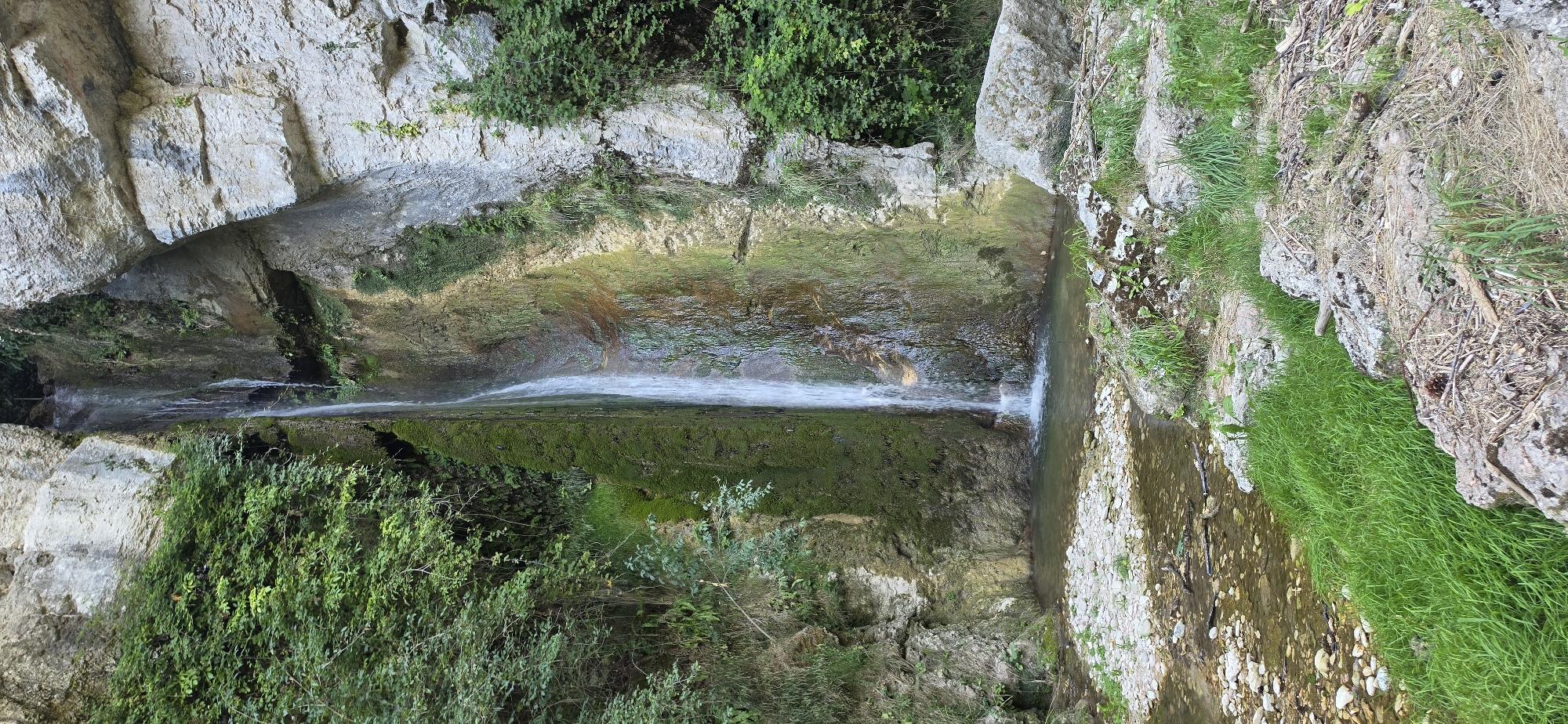 A vertical medium shot captures a narrow waterfall cascading down a moss-covered rock face within a natural canyon. Lush green foliage frames the left side of the waterfall, with dense bushes clinging to the rocky walls. The water flows into a shallow pool at the base, surrounded by a rocky creek bed dotted with white stones and a small amount of clear water. In the foreground, a patch of vibrant green grass grows next to a large boulder, with dry leaves and twigs scattered on the ground. The overall scene depicts a tranquil, natural environment. The location is identified as Fumane, Italy.